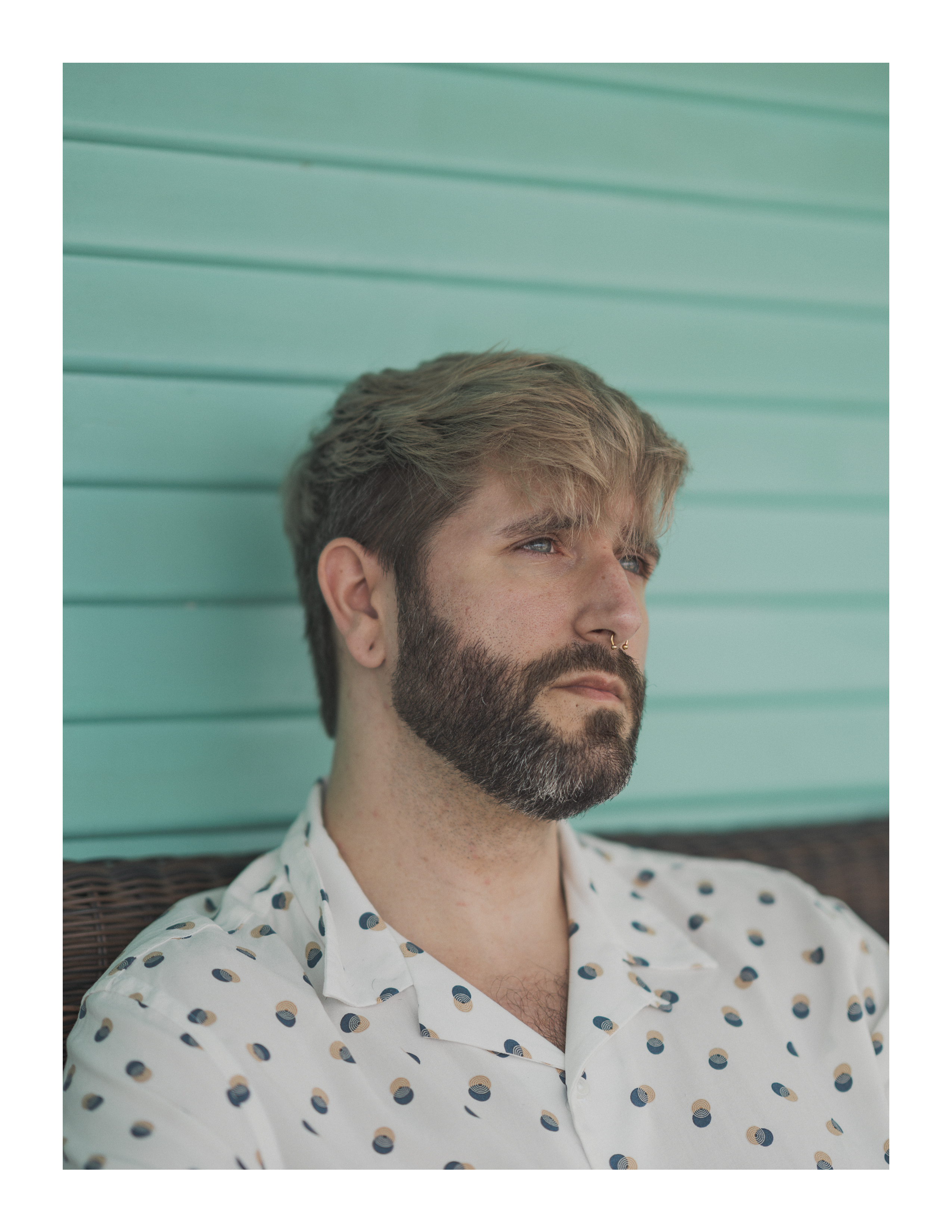 Editorial portrait of man in patterned shirt, seated against teal siding in soft natural light