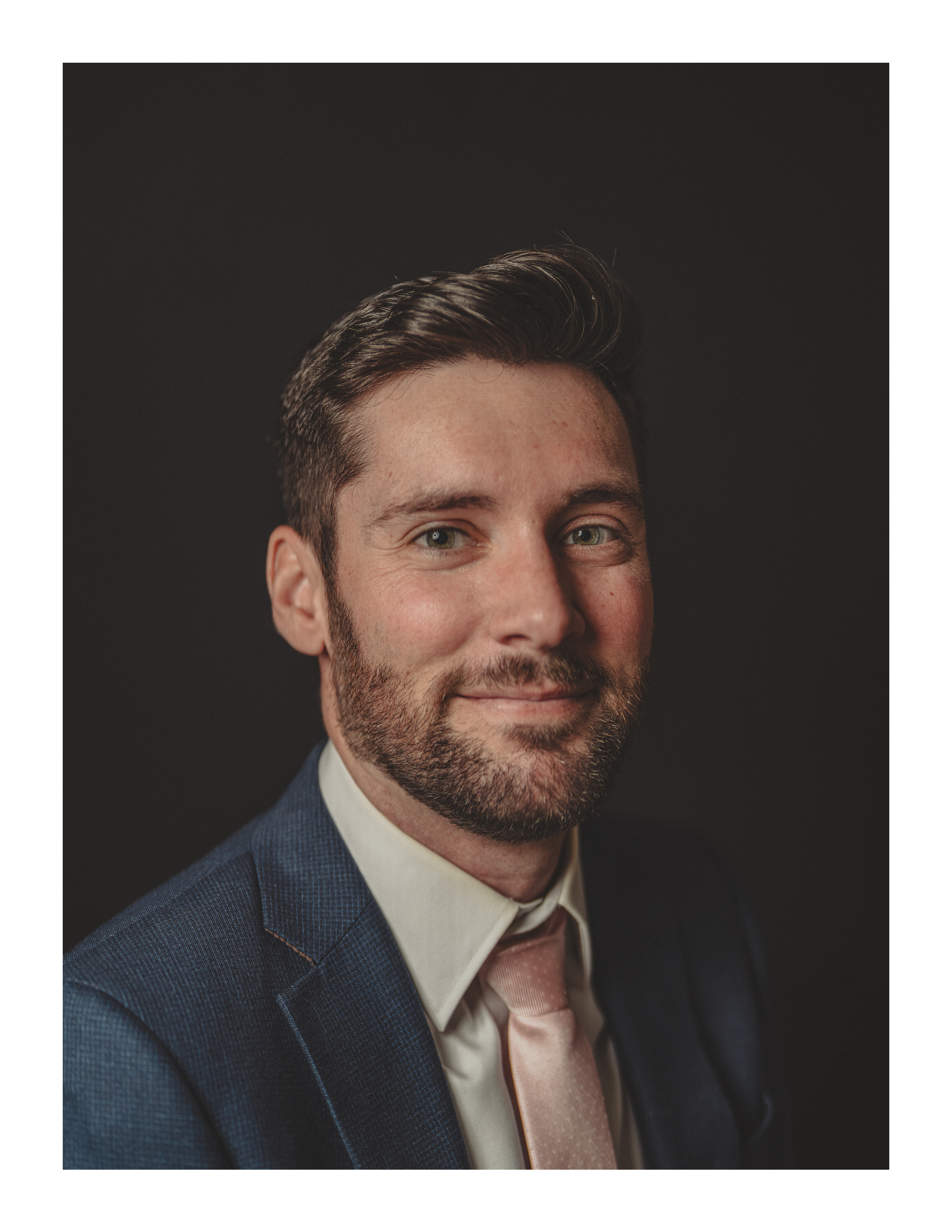 Studio portrait of man in blue suit and pale pink tie against black backdrop