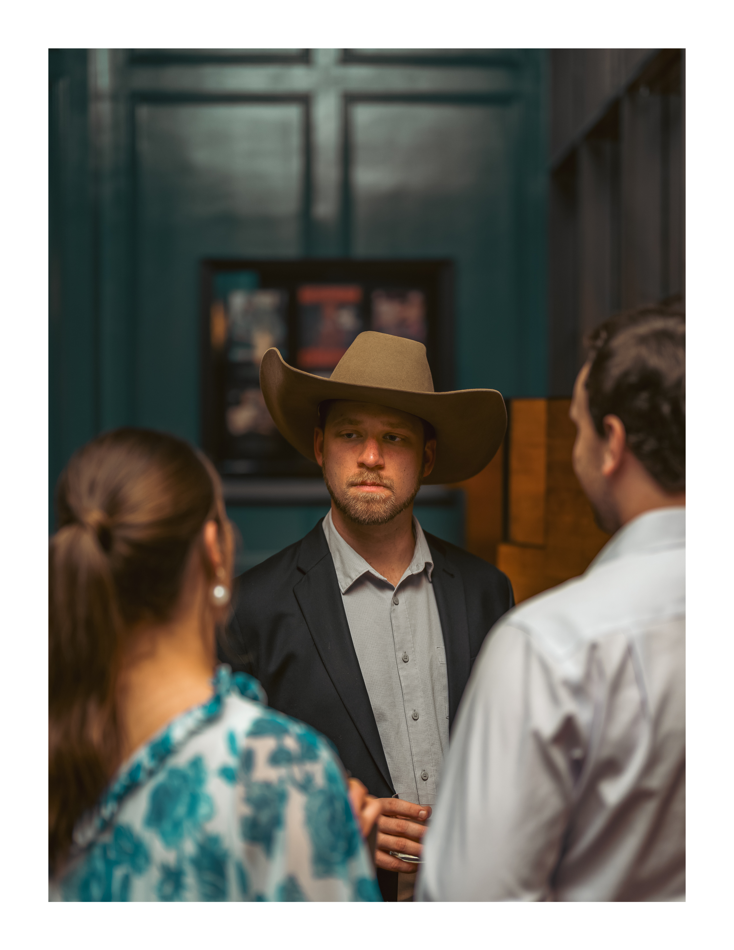 Man in cowboy hat and blazer mid-conversation in dim teal hallway with framed posters