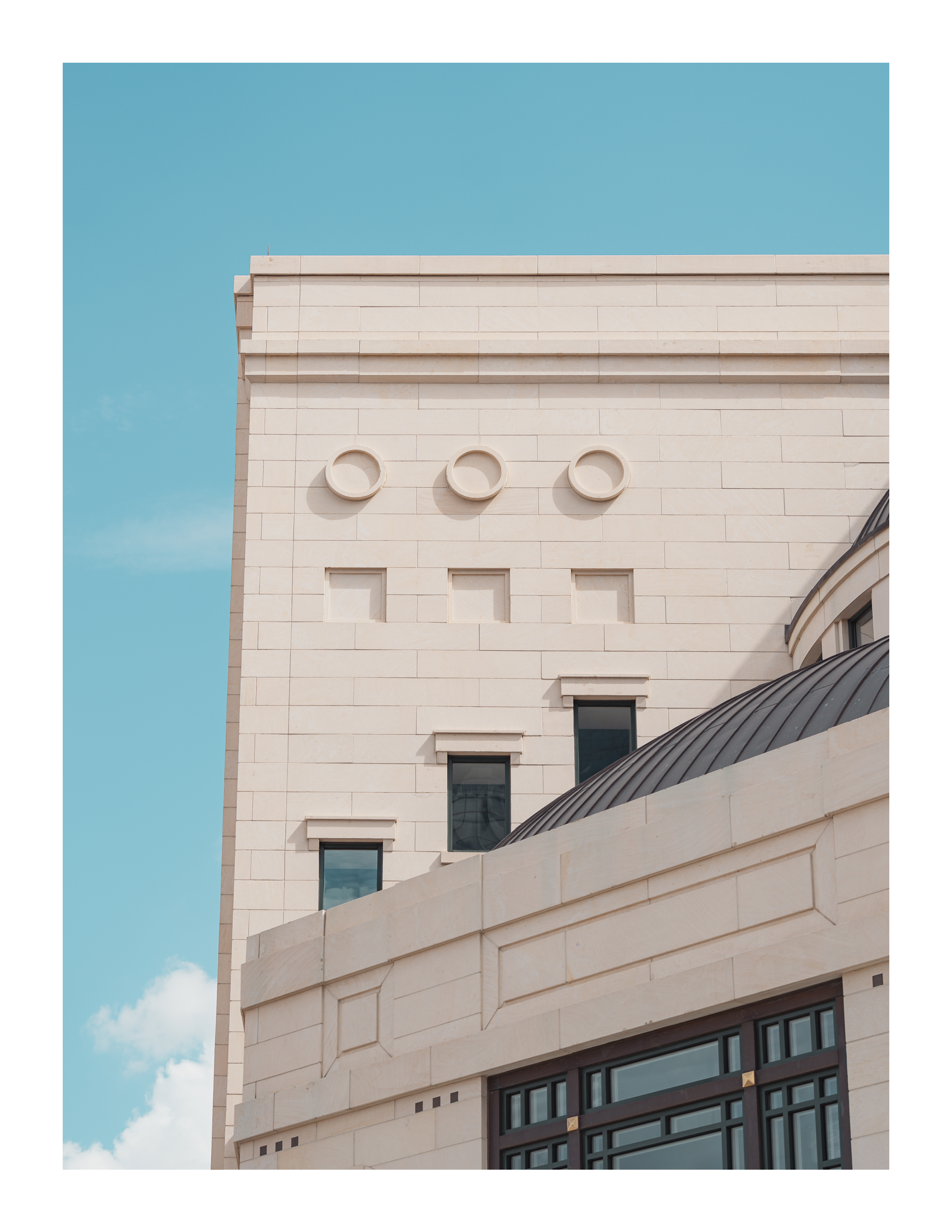 Cream-toned architectural façade with round windows, dome roof, and geometric detailing under blue sky