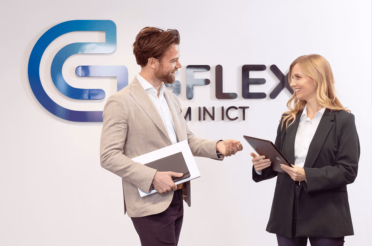 Een man in een licht colbert en een vrouw in een donker pak staan lachend met elkaar te praten voor een witte muur met het GFLEX logo. De man houdt een laptop en een notitieblok vast, en de vrouw heeft een tablet in haar handen.
