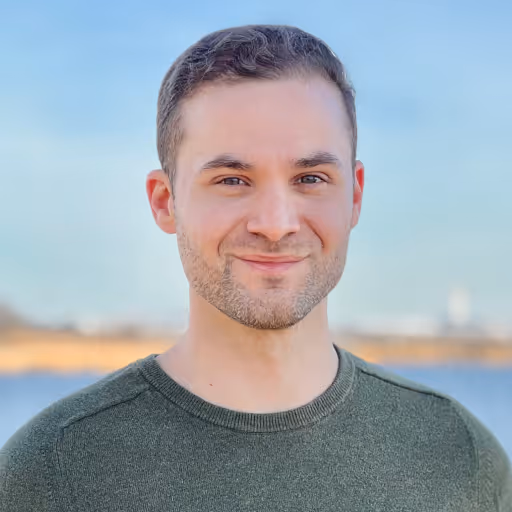 Smiling young man with short brown hair and trimmed beard wearing a green sweater, with a blurred outdoor background.