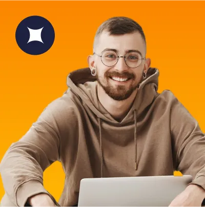 Smiling young man with glasses and earrings wearing a brown hoodie, sitting behind a laptop against an orange background.