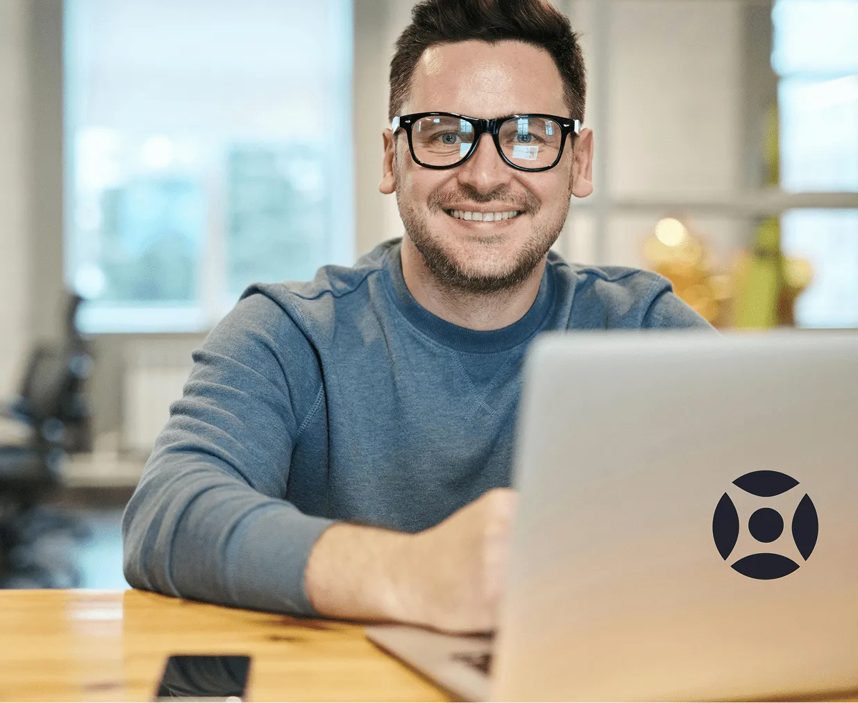 Smiling man with glasses working on a laptop at a wooden desk in a bright office.