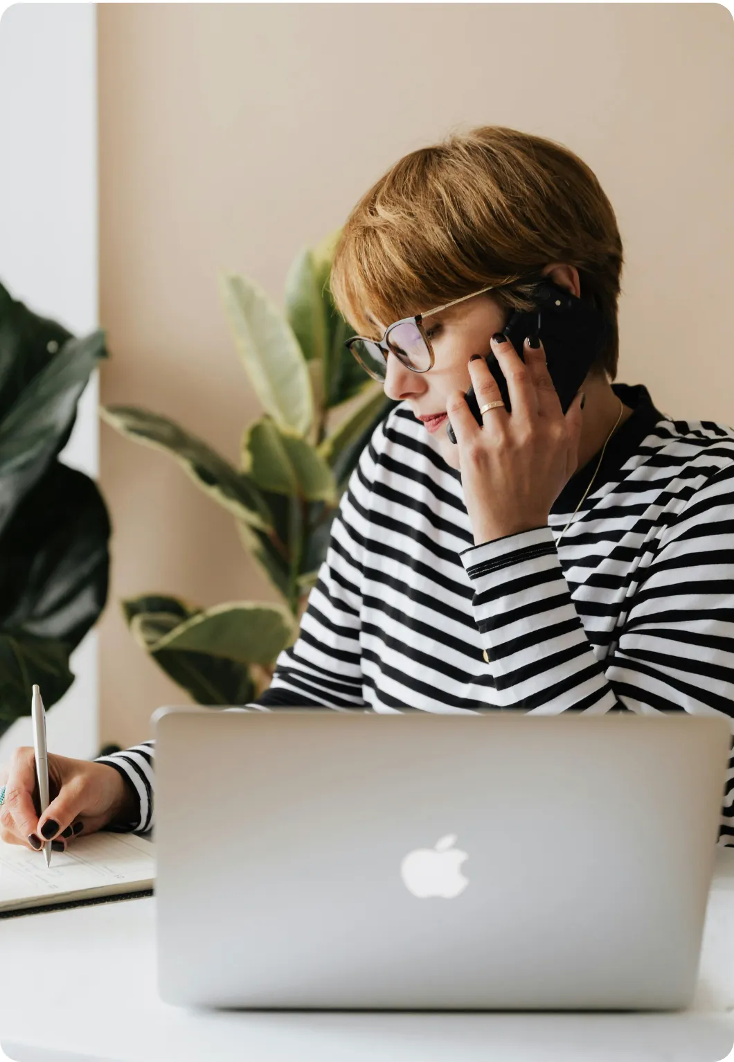 Person wearing glasses and a striped shirt talking on a smartphone while writing in a notebook behind a laptop.