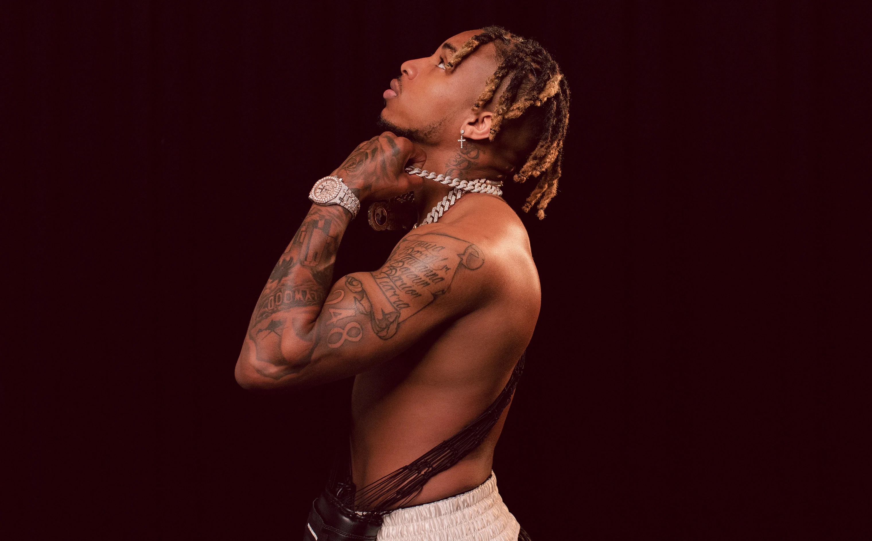 Shirtless man with tattoos and braided hair wearing chain jewelry against dark background