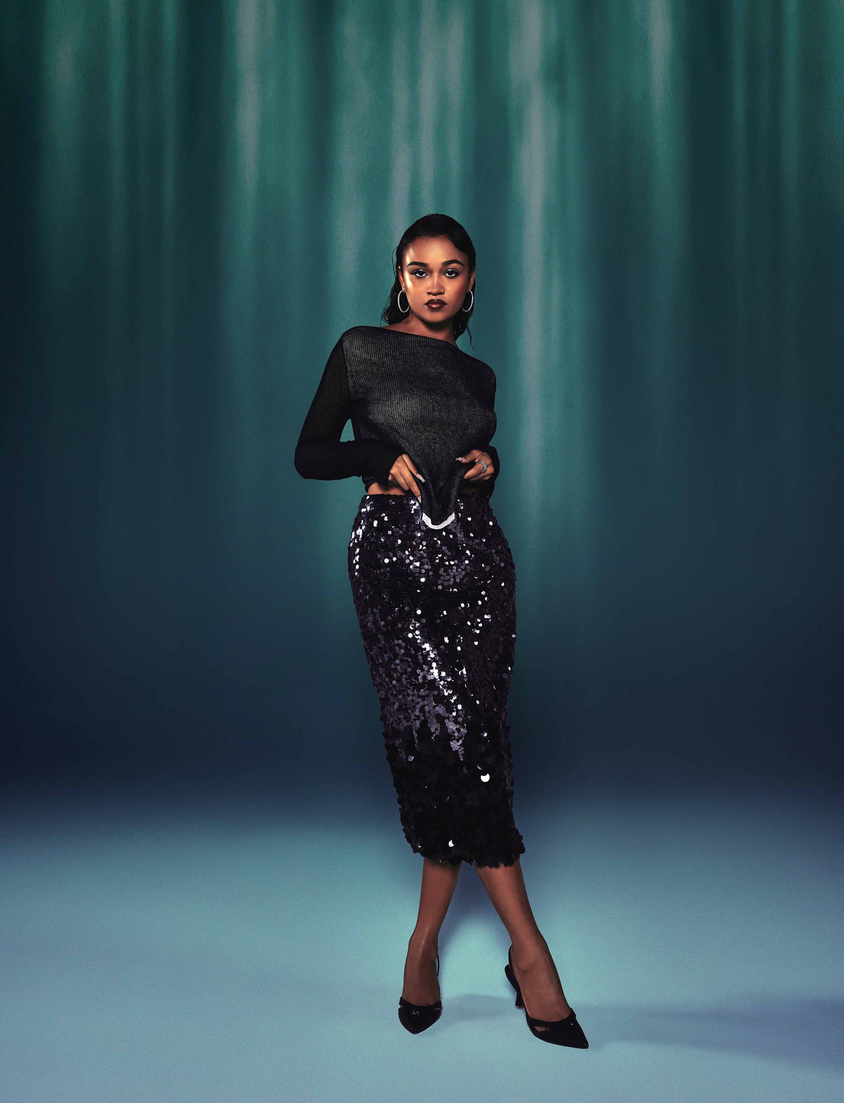 Woman in black knit top and sequined skirt poses against teal gradient background.