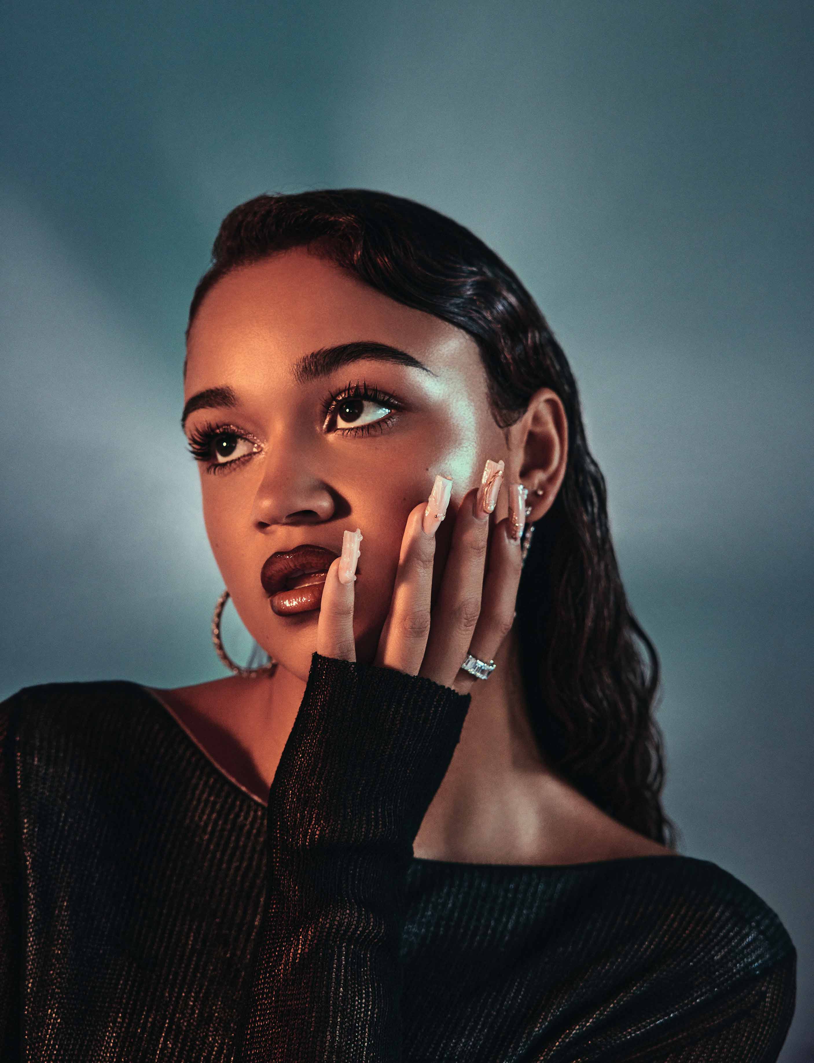 Woman with dark hair wearing black top and white acrylic nails poses against blue gradient background.