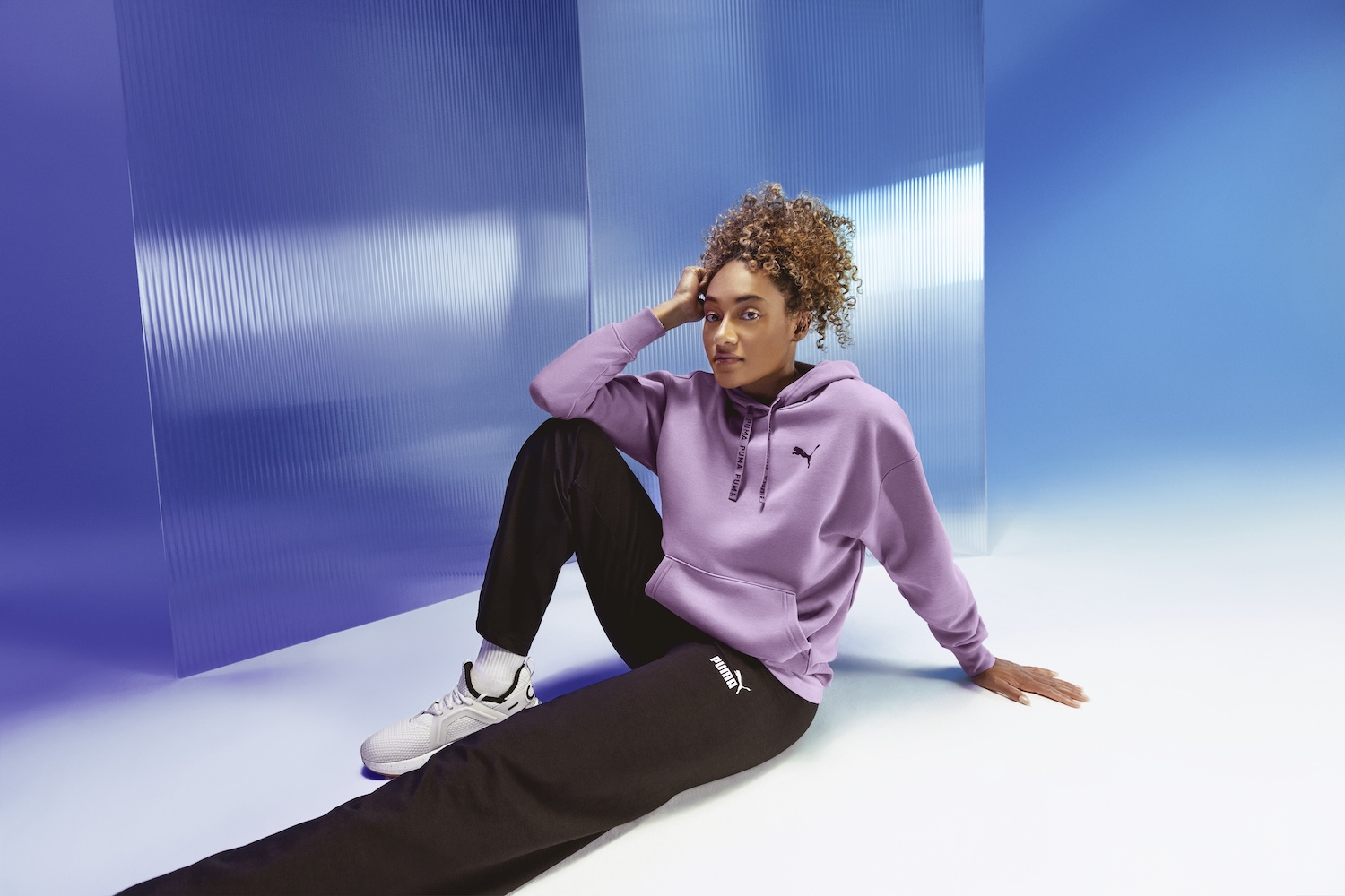 Woman in pink hoodie and black pants sits against blue wall backdrop, looking away thoughtfully.