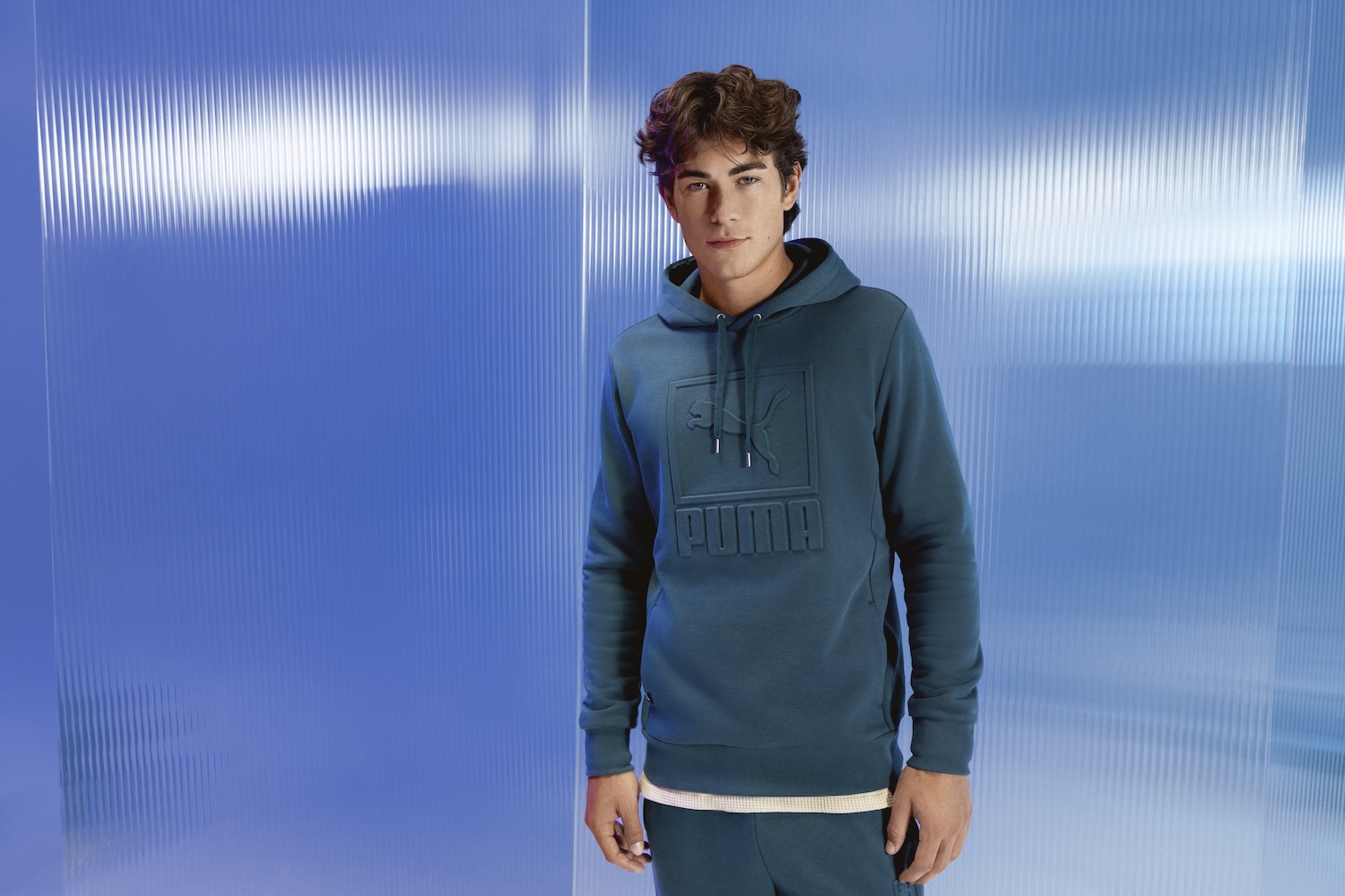 Young man wearing teal Puma hoodie against blue striped wall background