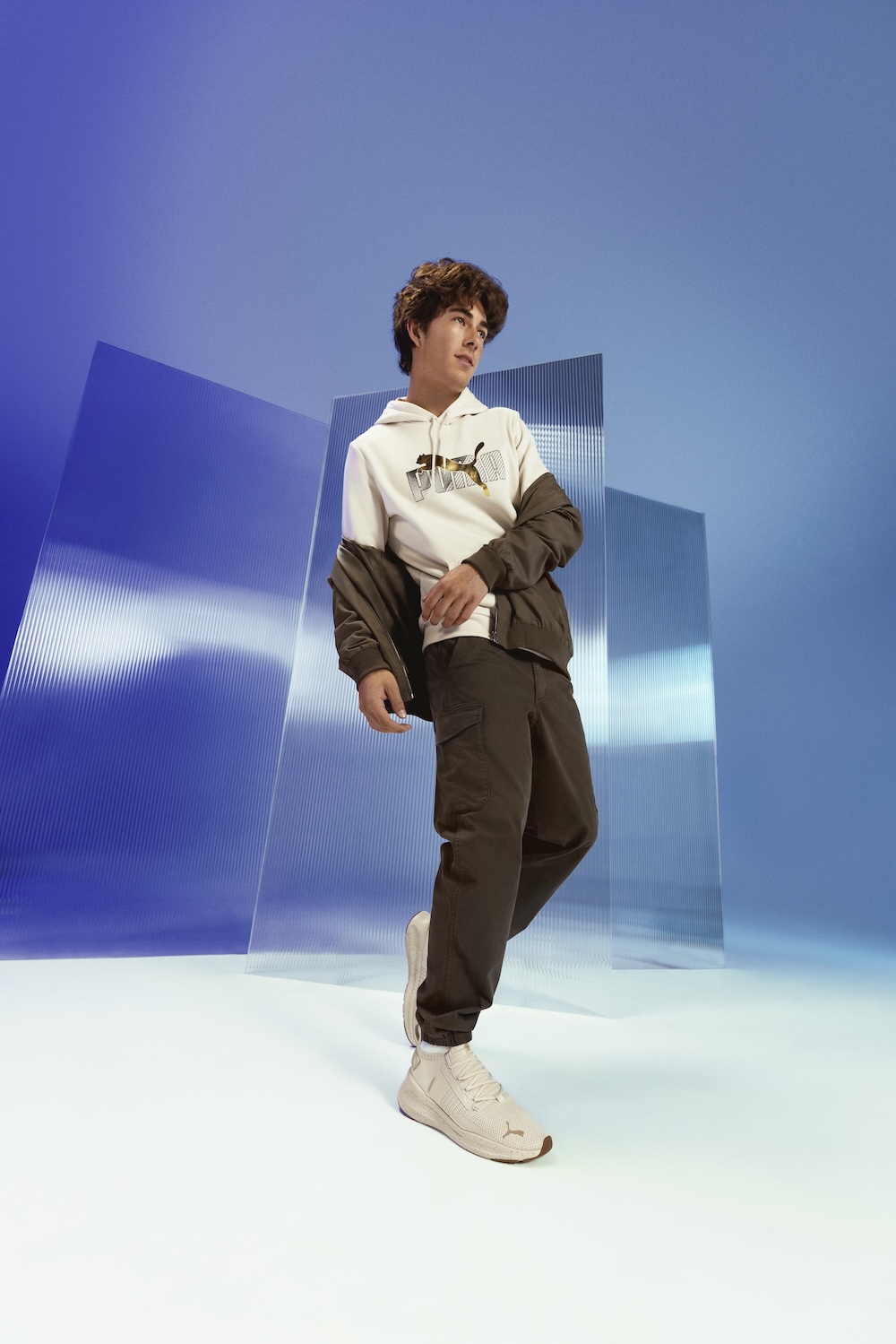 Young man in cream hoodie and brown jacket posing against blue geometric backdrop