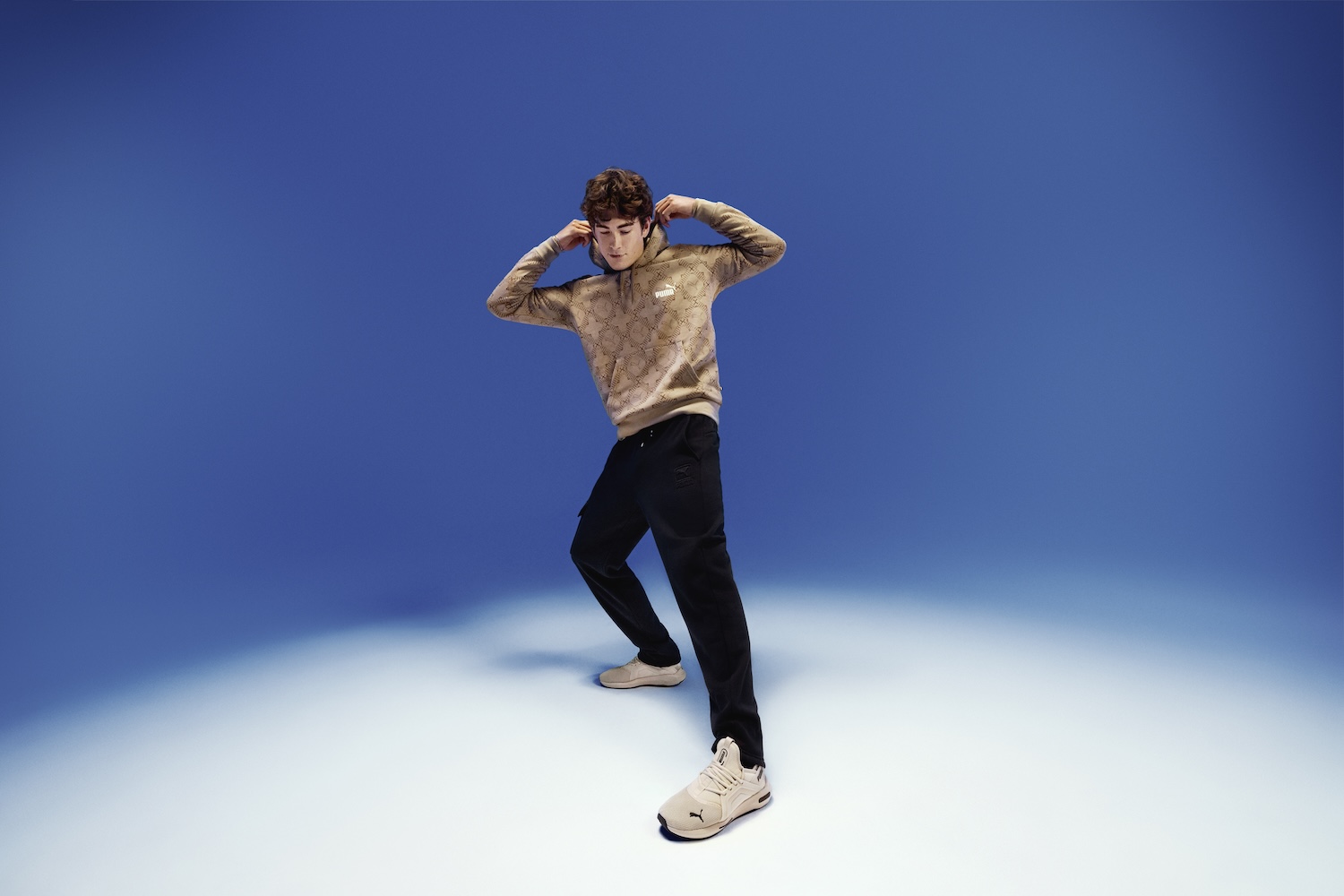 Young person in beige jacket and black pants posing dynamically against blue background
