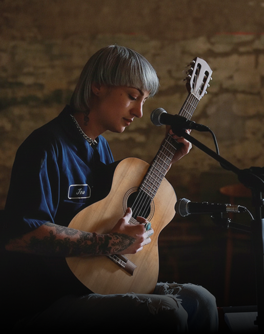 Música con cabello corto tocando una guitarra acústica frente a un micrófono en un ambiente íntimo.