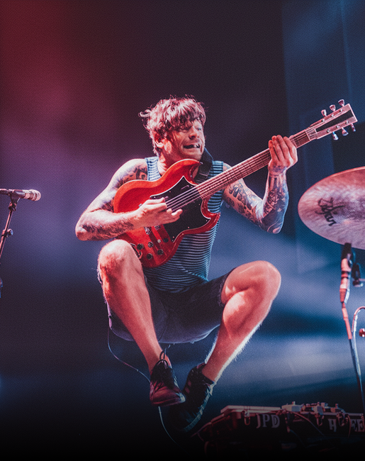 Músico tatuado saltando en el aire mientras toca una guitarra eléctrica roja en un escenario iluminado.