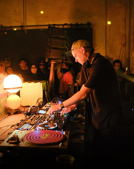 DJ con gorra y auriculares operando una mesa de mezclas en un evento nocturno con iluminación cálida.