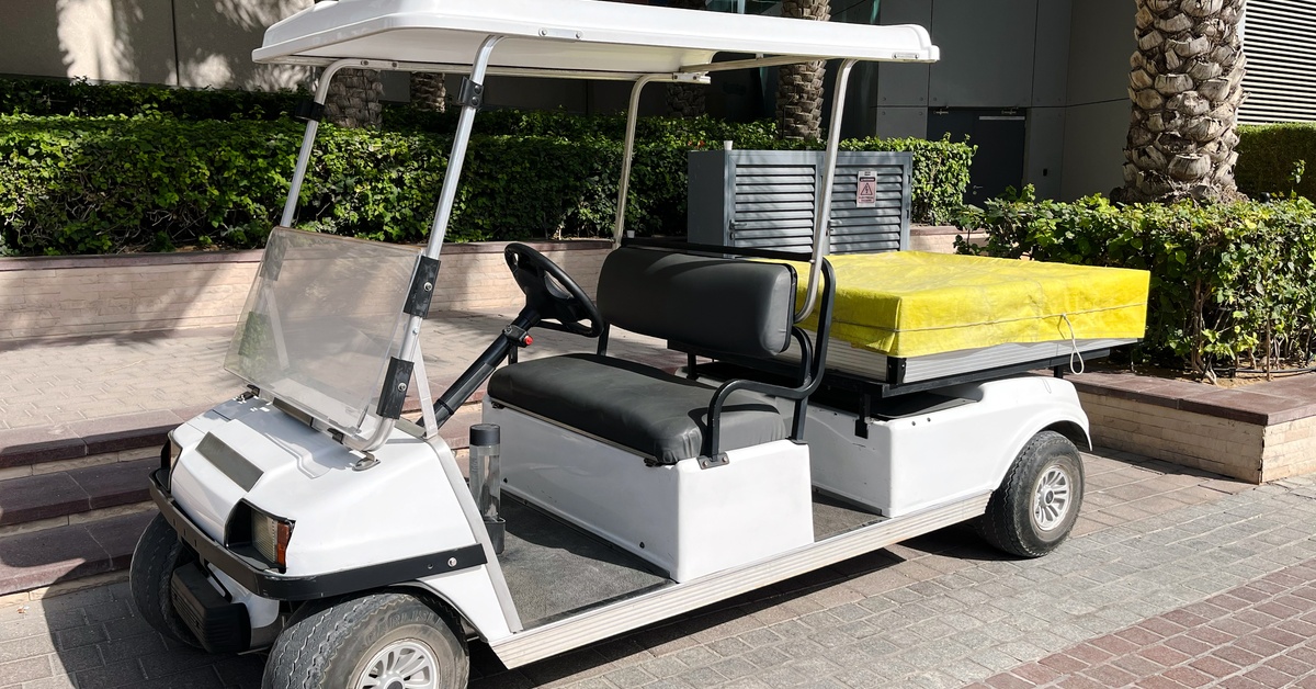 A white golf cart with black seats, a windshield, and a trunk attachment covered with a yellow tarp parked on the street.
