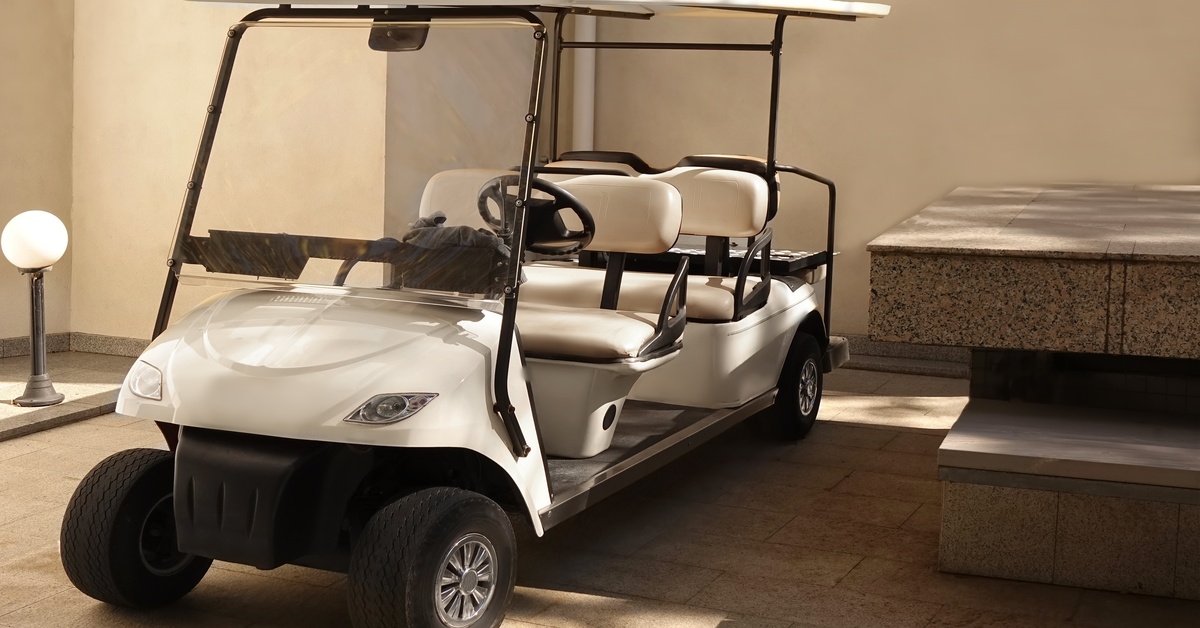 White golf cart with six seats, a full windshield, and a complete roof, parked between a large concrete bench and a lamp.