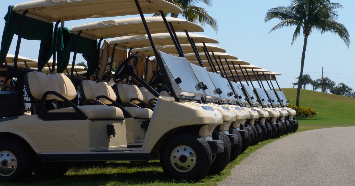 Multiple new beige golf carts with two seats parked next to each other over grass aligned with a pavement train.
