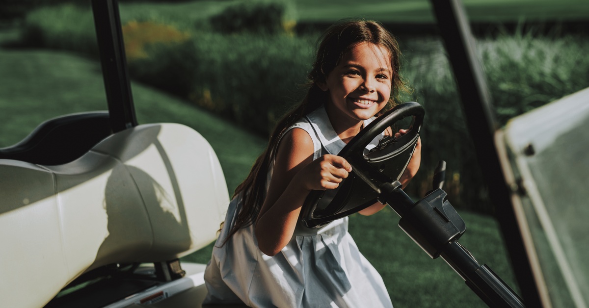 A young girl wearing a dress sits behind the wheel of a golf cart parked in a grassy field on a sunny day.