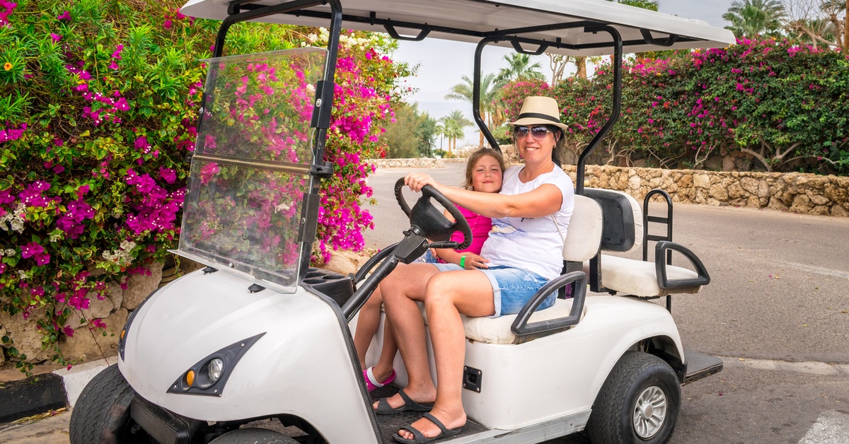 A woman and a young girl sit next to each other on a golf cart while riding along a concrete road lined with bougainvilleas.