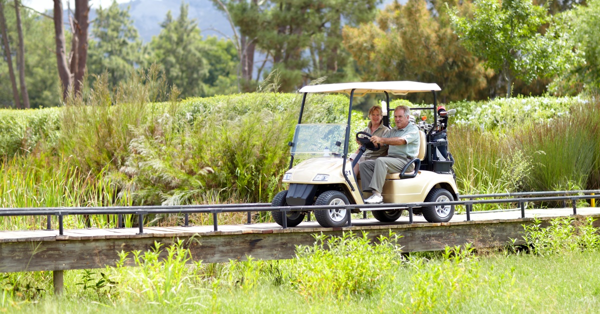 An older couple riding on a golf cart. They are driving over a wooden path bridge surrounded by green bushes and trees.