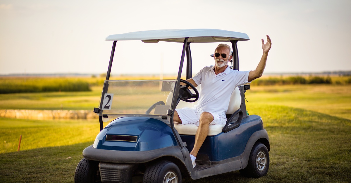 A senior man in a white polo shirt and white shorts waving while sitting behind the wheel of a golf cart outdoors.