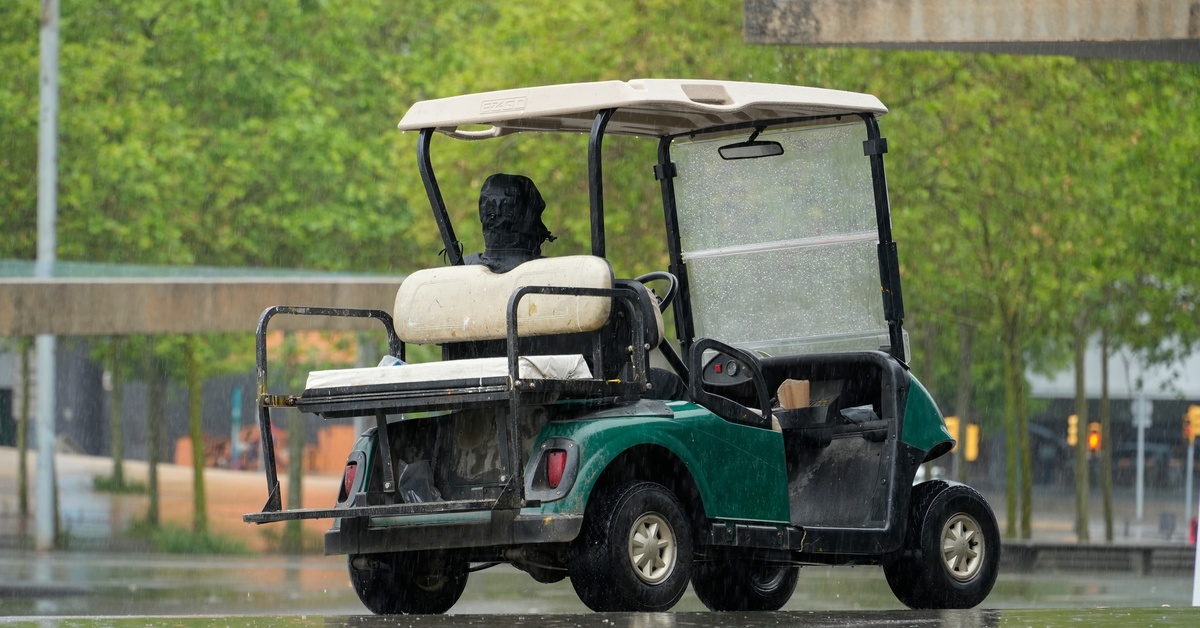 A person wearing a black jacket with a hoodie sits in a parked golf cart. It is raining, and there are trees in the background.