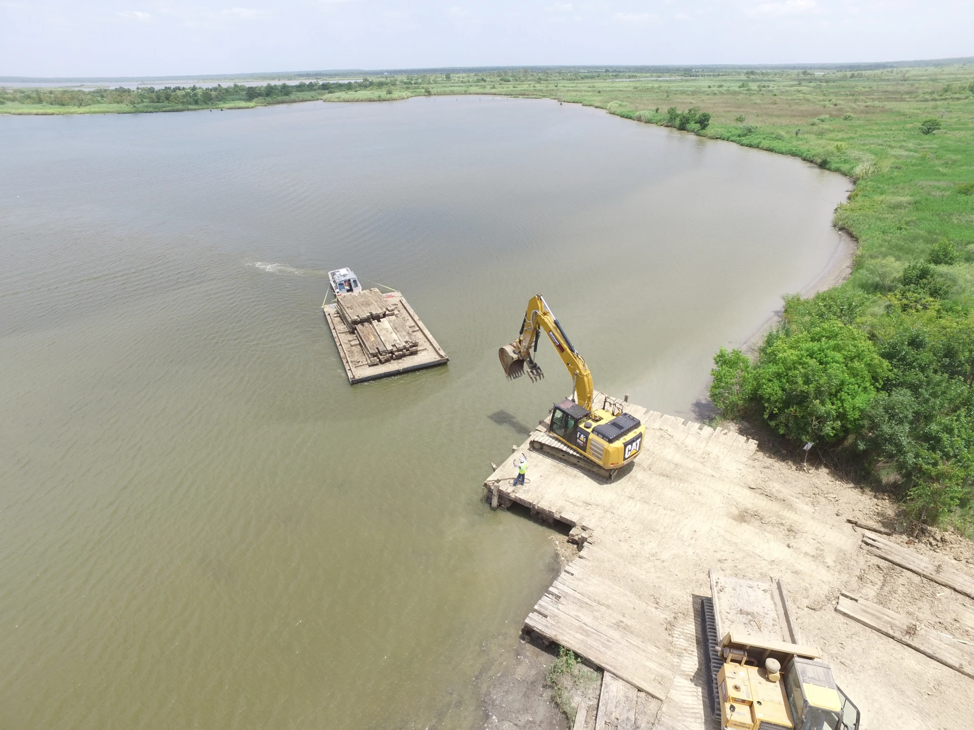 An aerial view of a construction site near a body of water, featuring an excavator on land and a barge on the water. The surrounding area includes green fields and a clear sky.