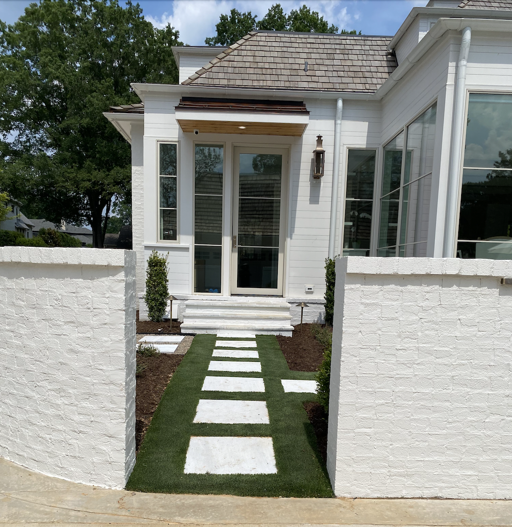 Artificial turf is used around square pavers that lead to a home’s side door. 