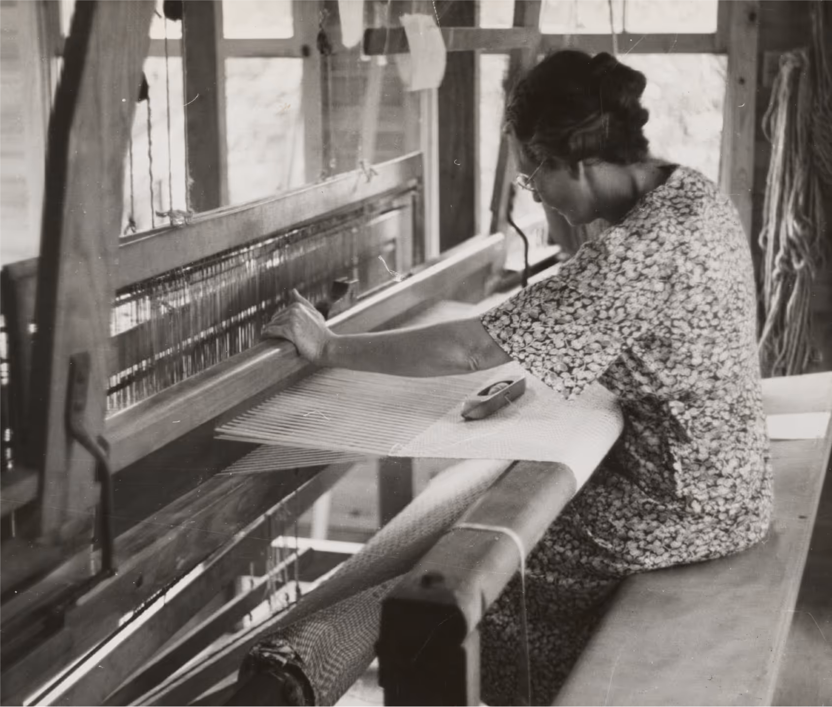 Black and white photograph of a woman weaving by hand on a traditional wooden loom, highlighting the craftsmanship behind textile making.