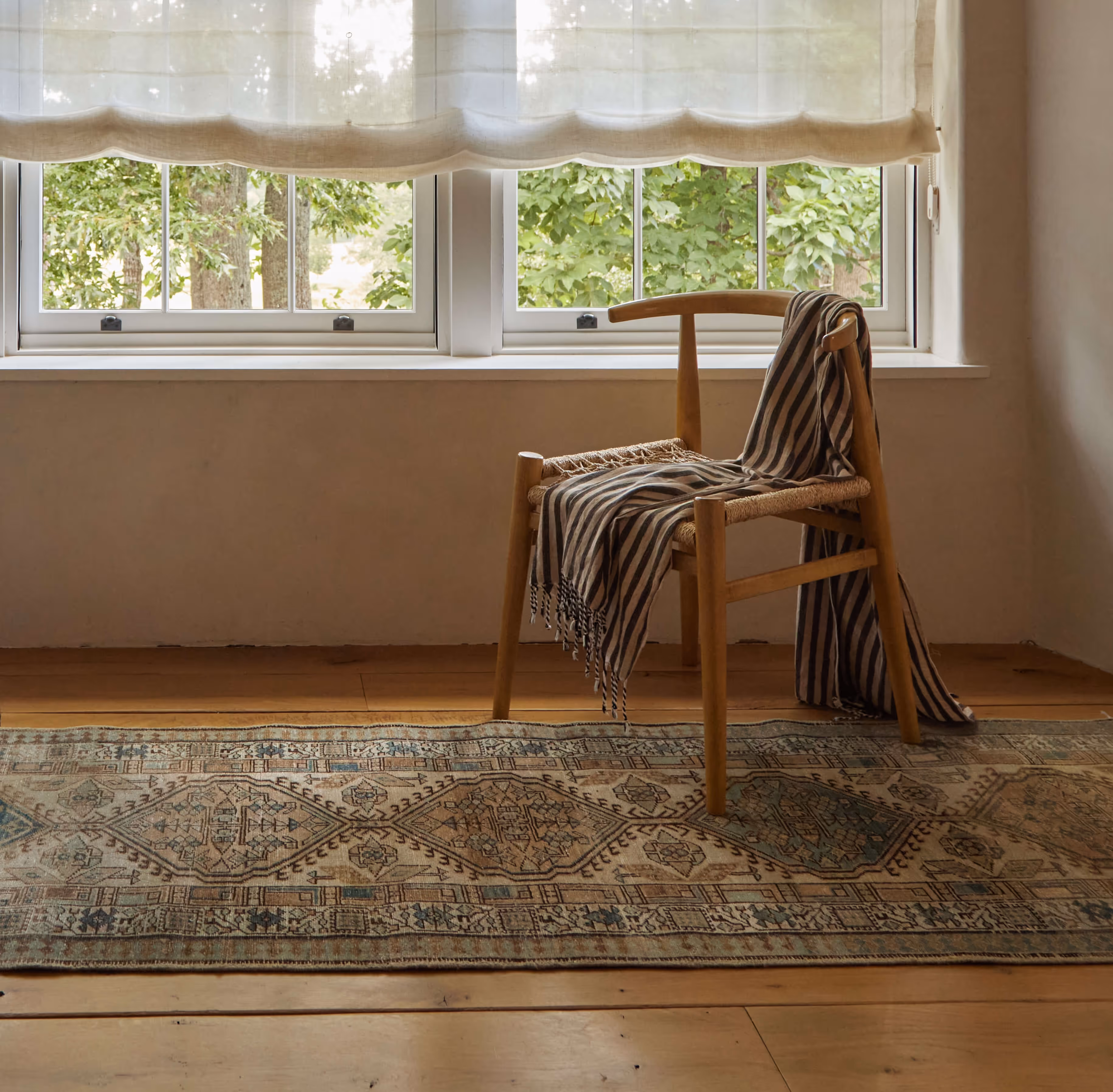 A quiet corner featuring a handwoven Moattar runner layered over wide plank floors, with a simple wooden chair draped in stripes, basking in the soft light of a shaded window.