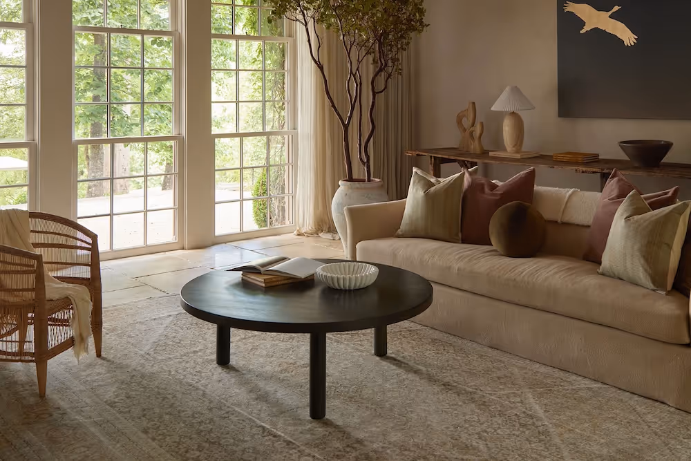 A calming living room with expansive windows, earthy tones, refined decor, and a subtly patterned rug creating an atmosphere of understated luxury.