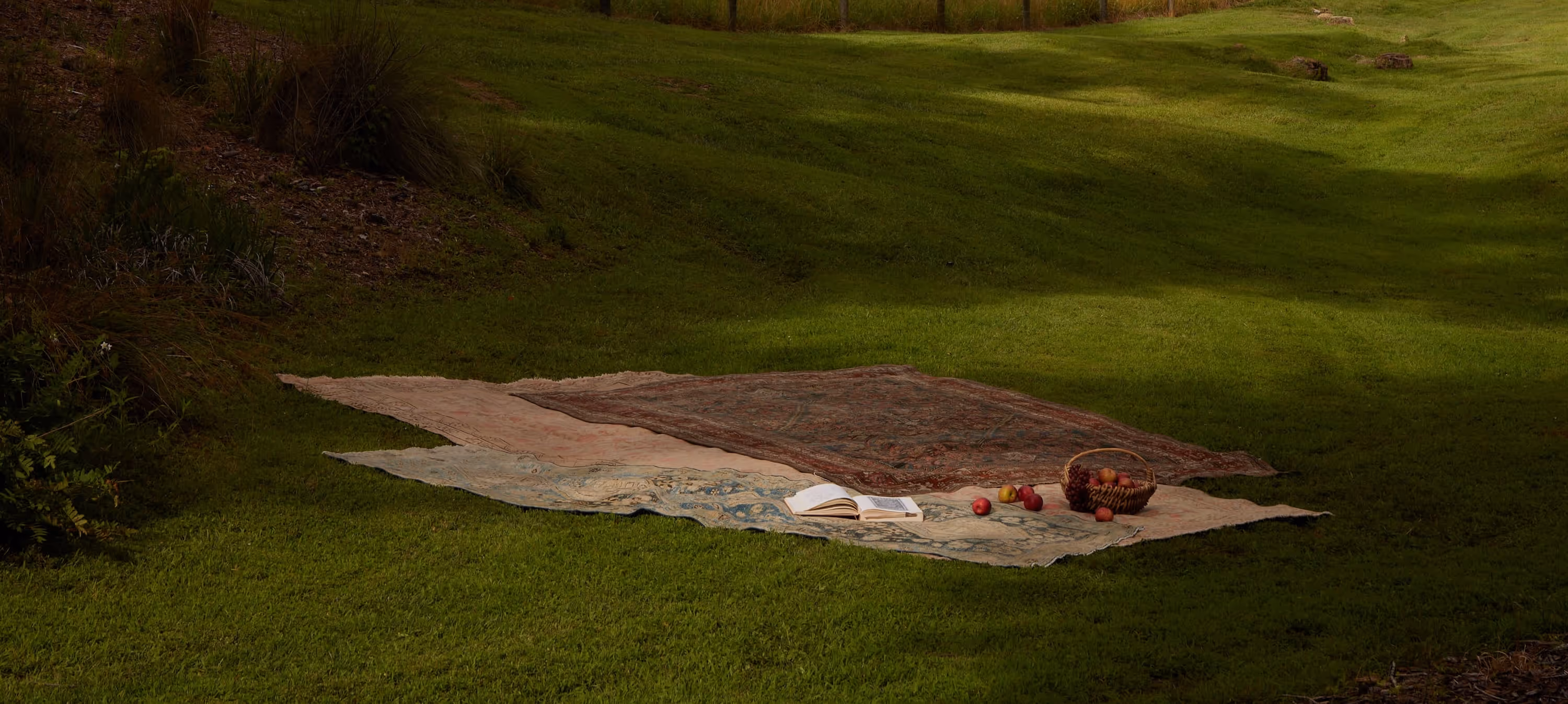 A tranquil outdoor picnic scene featuring layered vintage rugs on lush grass, complemented by fresh apples, an open book, and dappled sunlight, evoking timeless elegance and serenity.