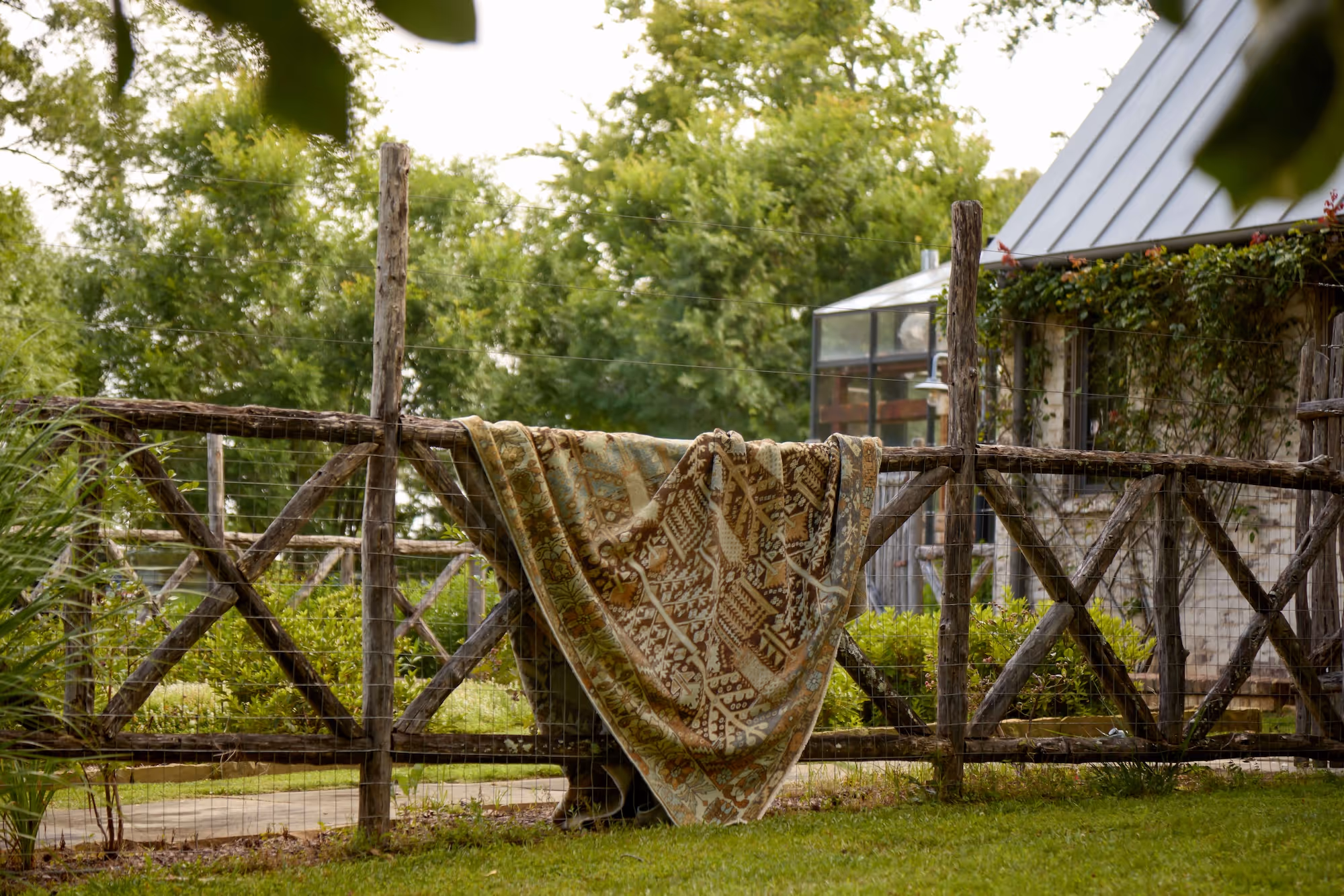 A vintage rug draped over a rustic wooden fence in a lush garden, capturing the timeless charm of Moattar’s outdoor styling.