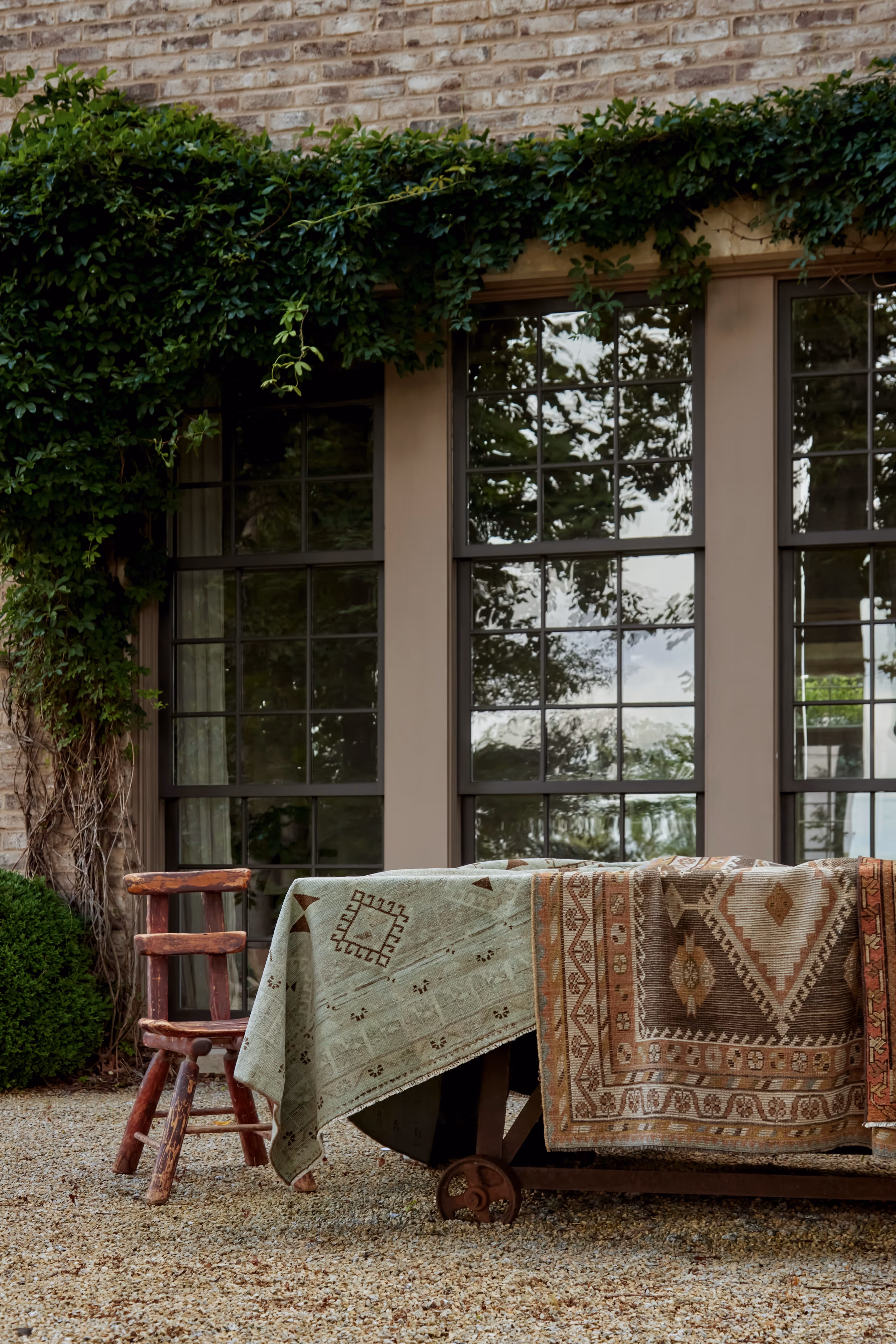 An inviting outdoor scene featuring a collection of artisanal rugs draped gracefully over a vintage table, framed by lush greenery and classic brick architecture.