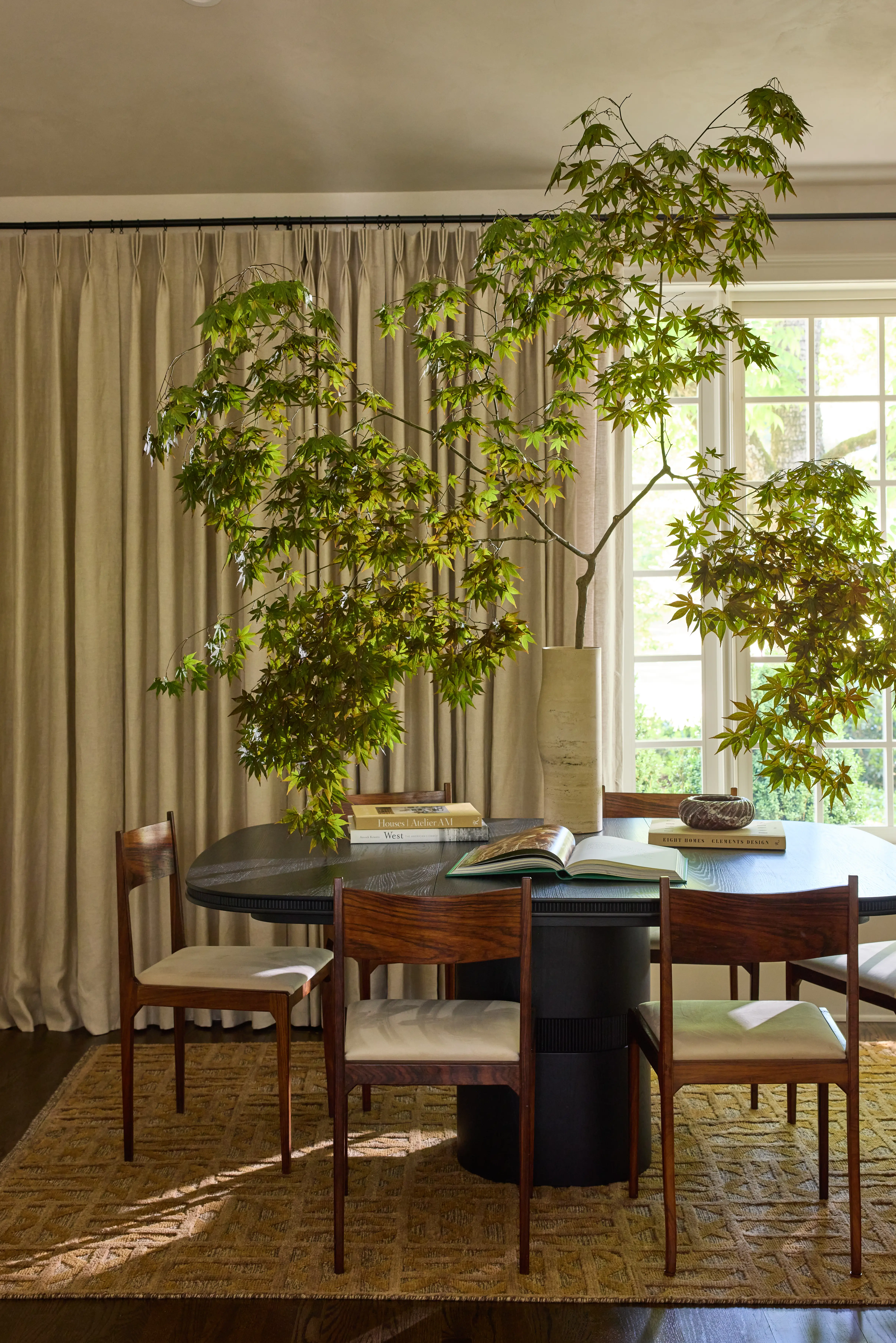 Dining table with six chairs, a large vase with green branches, books, and an open book by a window.