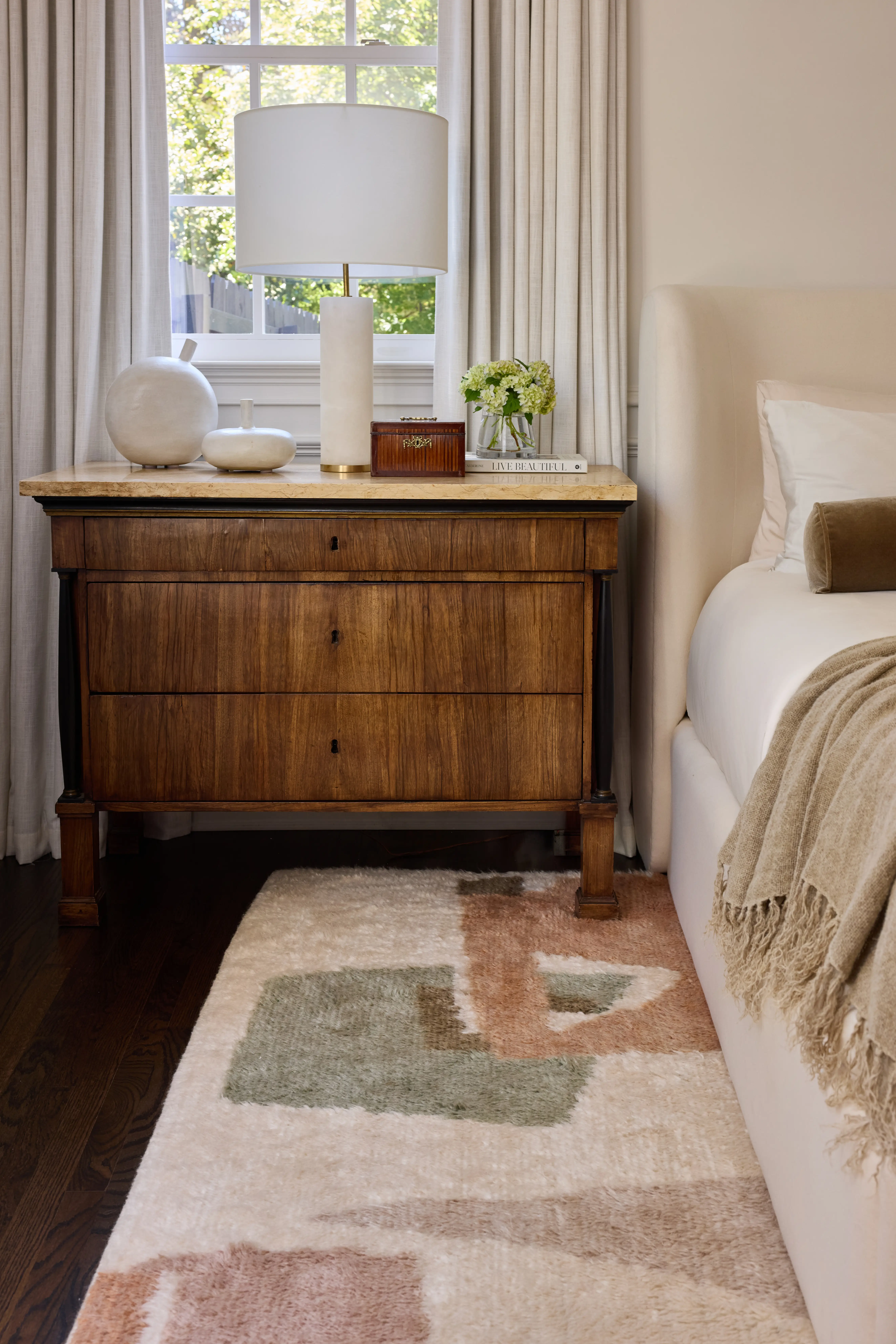 Wooden nightstand with white lamp, ceramic decor, small box, flowers, and book beside a cream upholstered bed.