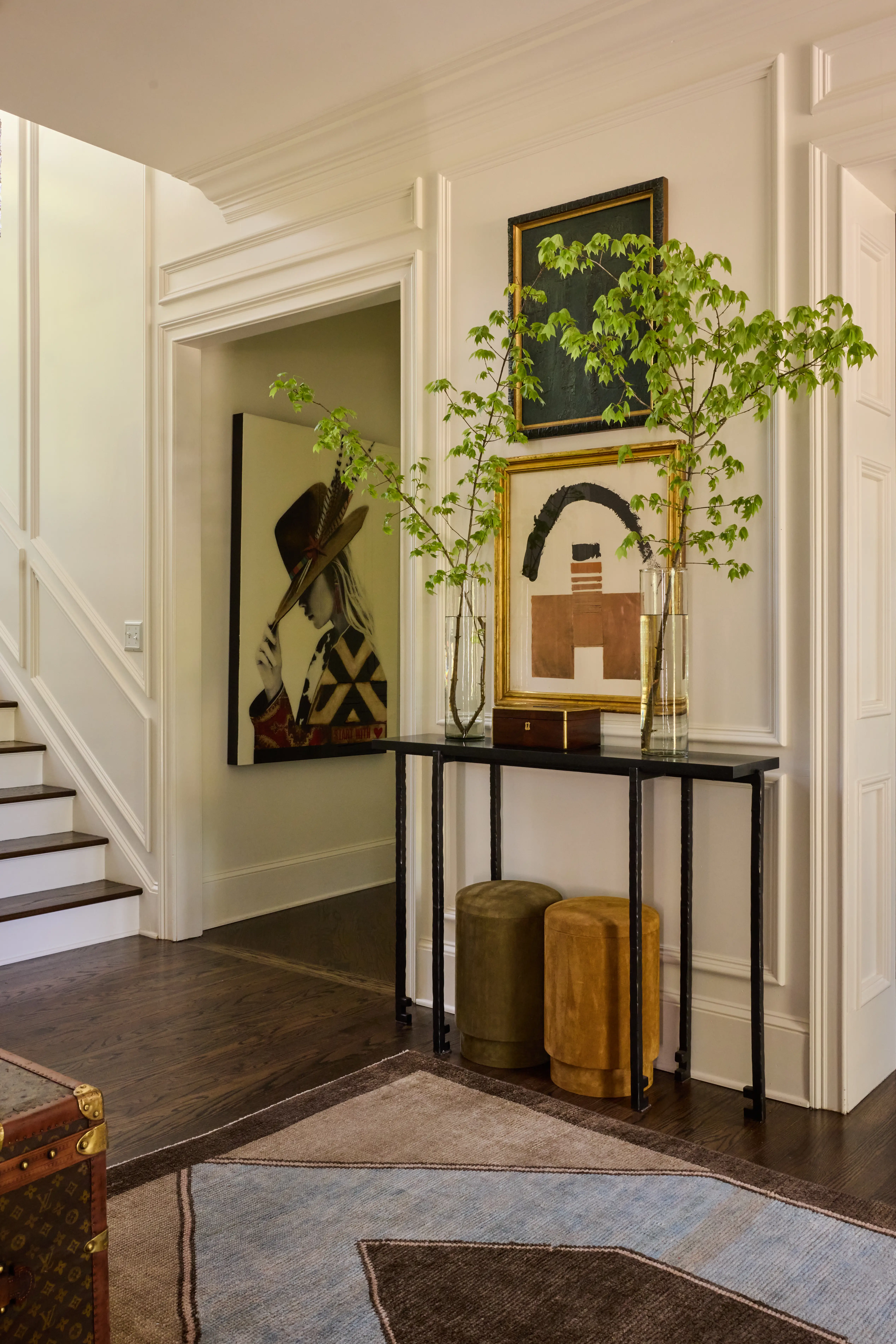 Entryway with black metal console table, two vases with branches, framed art, stools, and staircase.