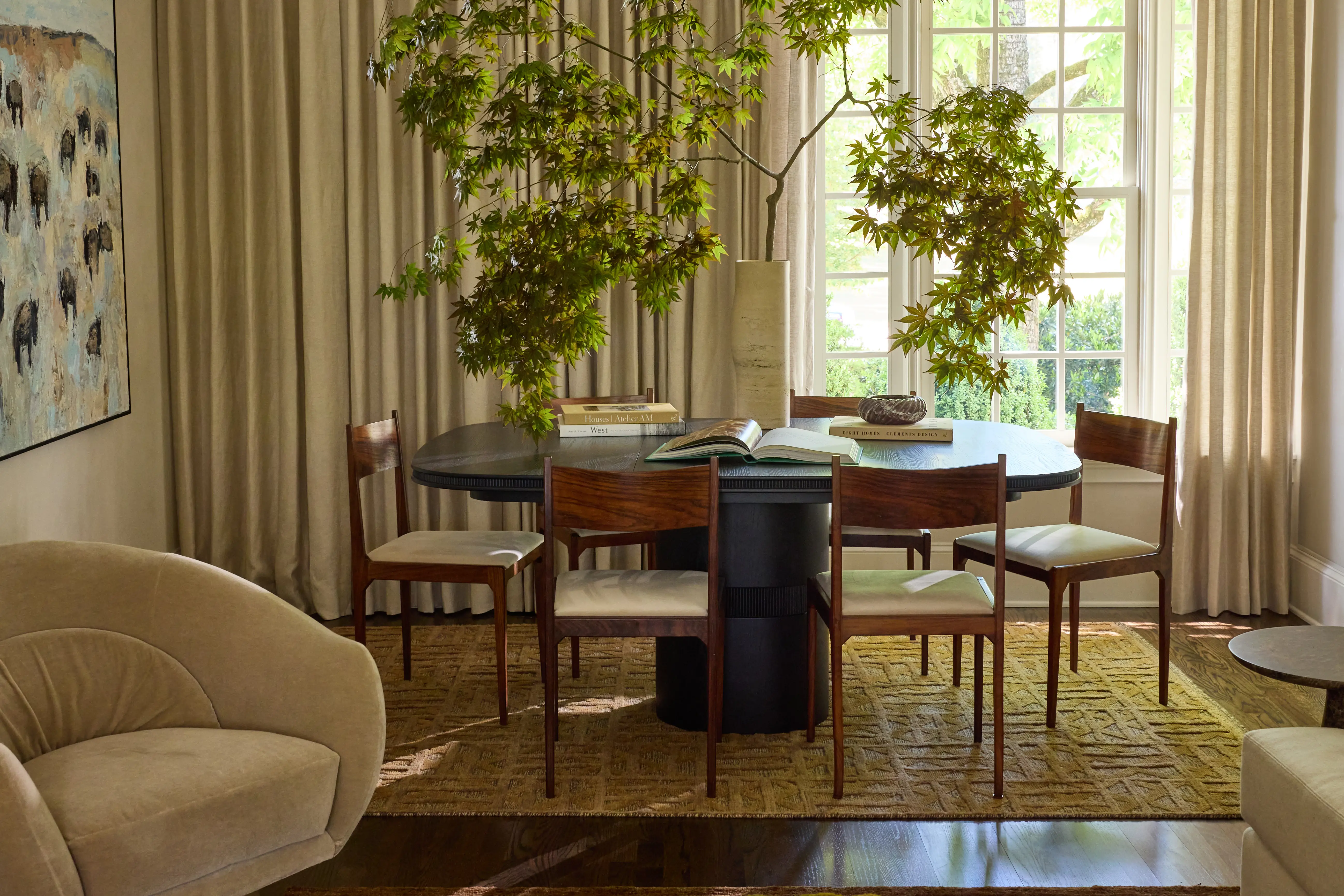 Dining room with a round black table, six wooden chairs, large vase with green branches, and beige curtains.