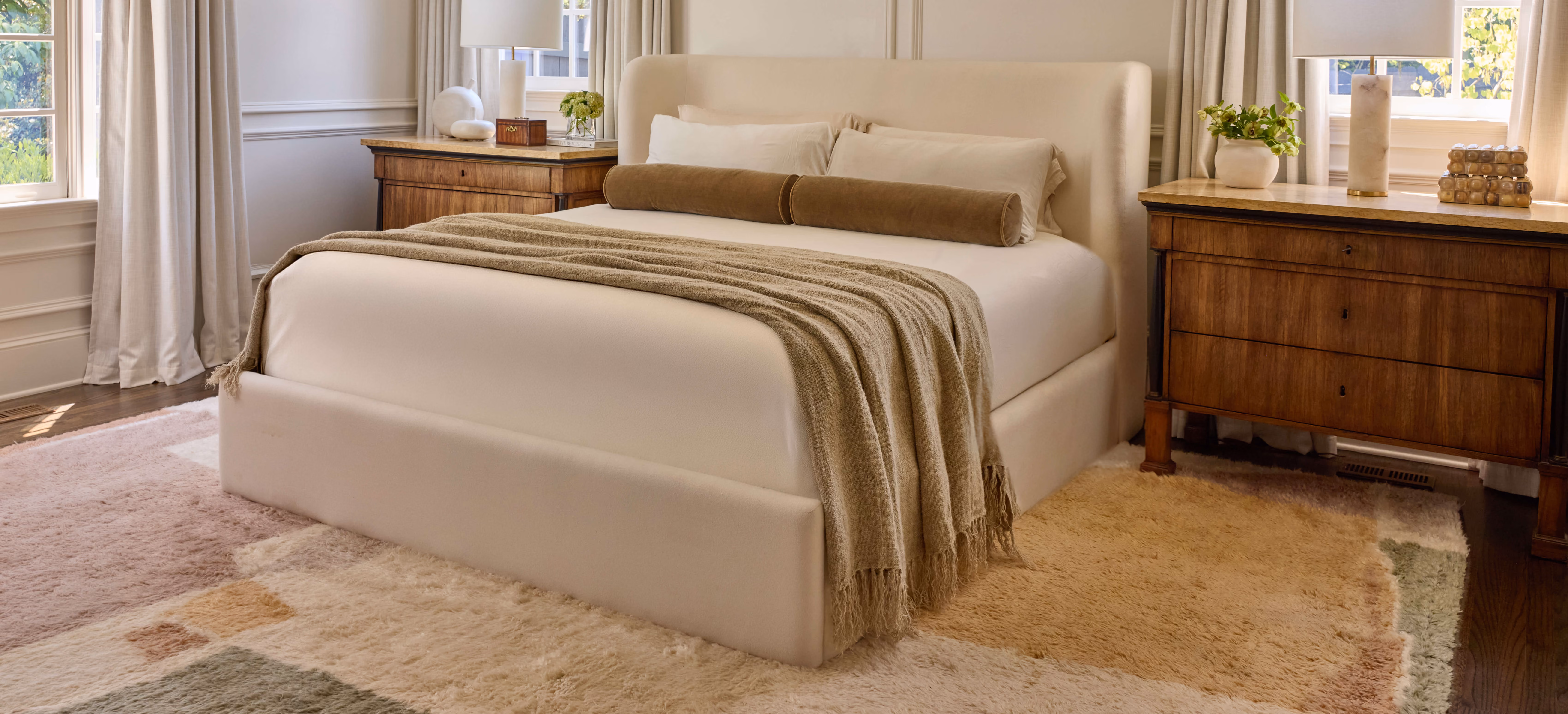 Beige upholstered bed with pillows and throw blanket between wooden nightstands in bright bedroom.