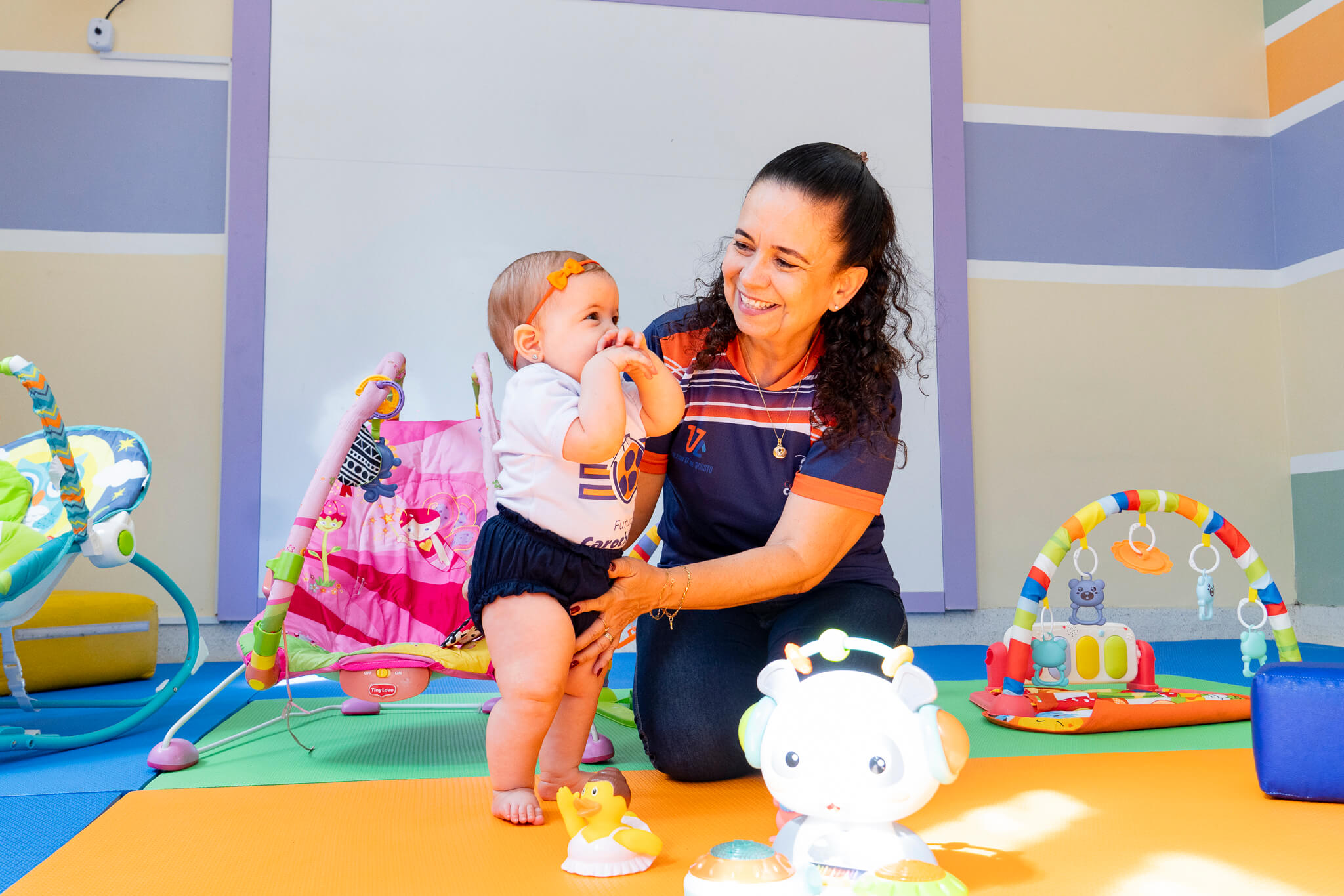 Professora do Centro Escolar Carochinha interagindo com bebê no berçário da escola na Zona Norte de Recife, em um ambiente colorido e seguro com brinquedos educativos.