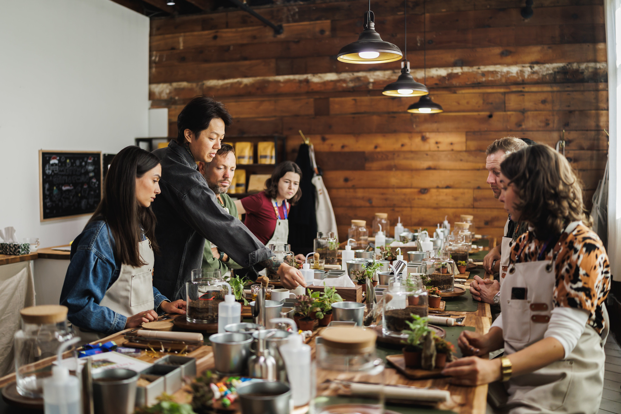 Attendees in aprons building glass terrariums at a long wooden workbench lined with plants and tools.