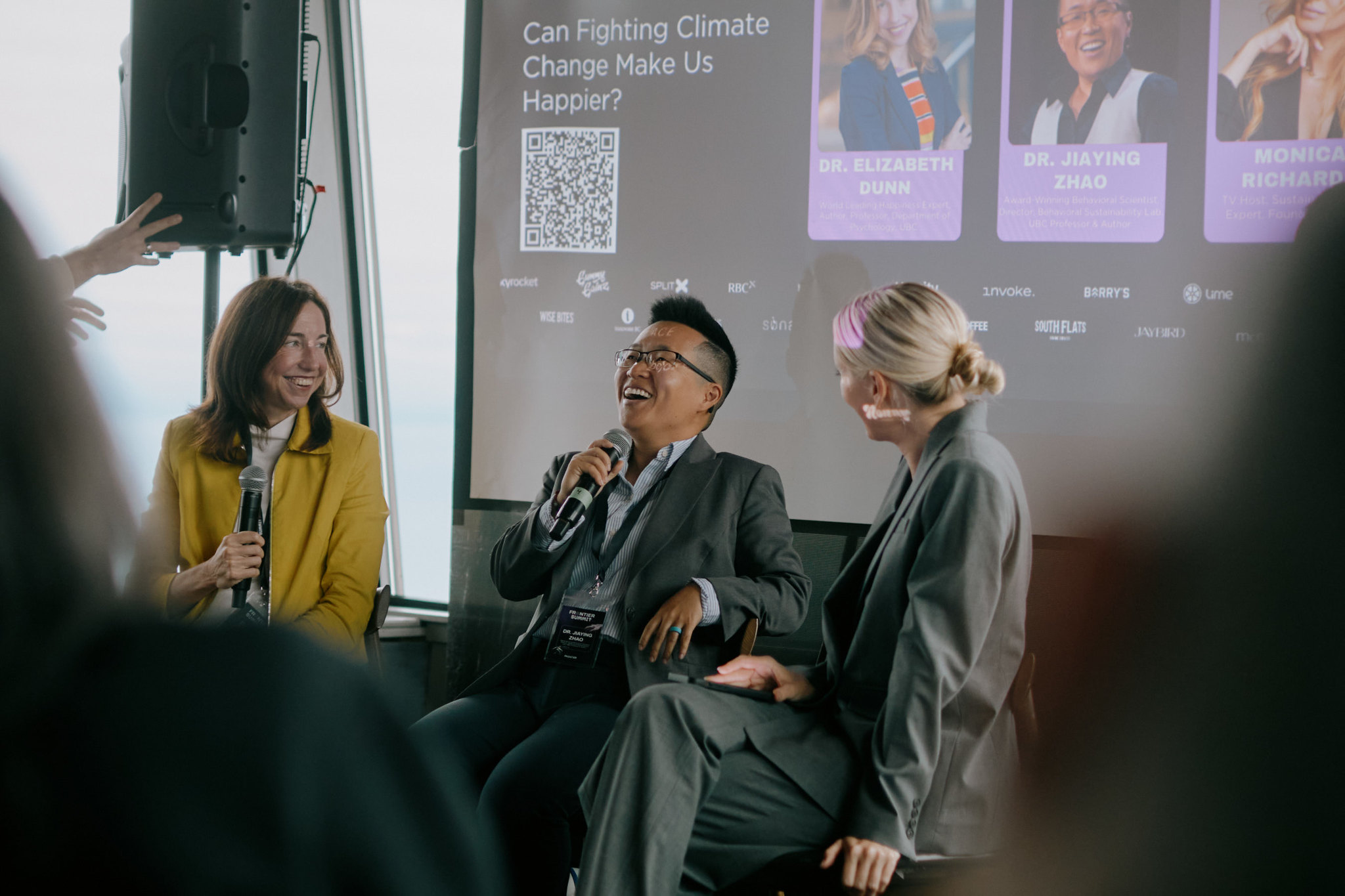 Three panellists laugh together on stage beneath a screen reading Can Fighting Climate Change Make Us Happier?