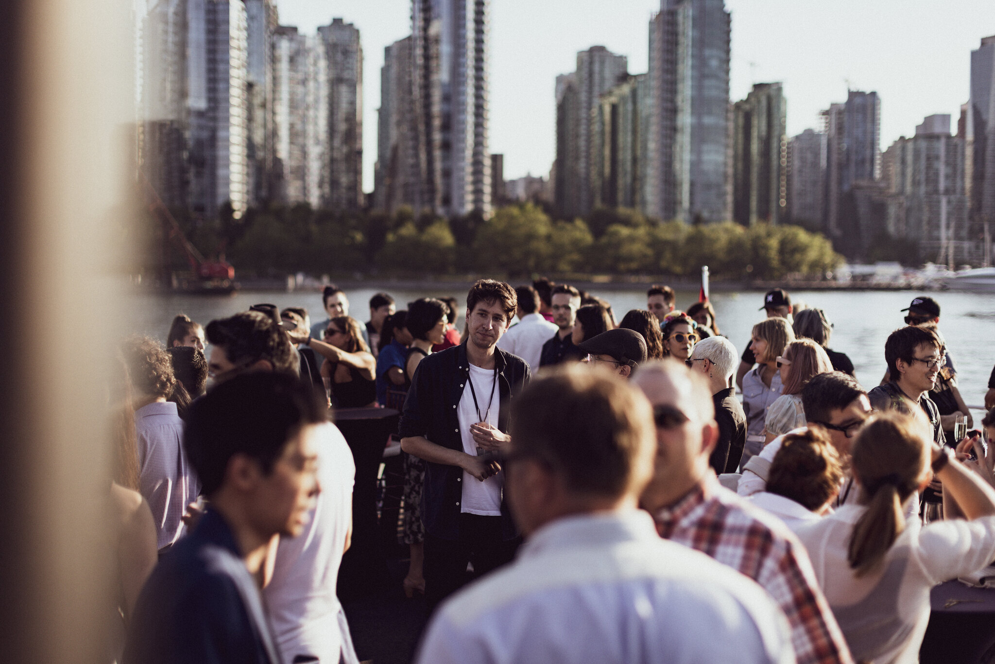 Guests mingling with drinks at an outdoor waterfront reception, with Vancouver's downtown towers rising across the water.