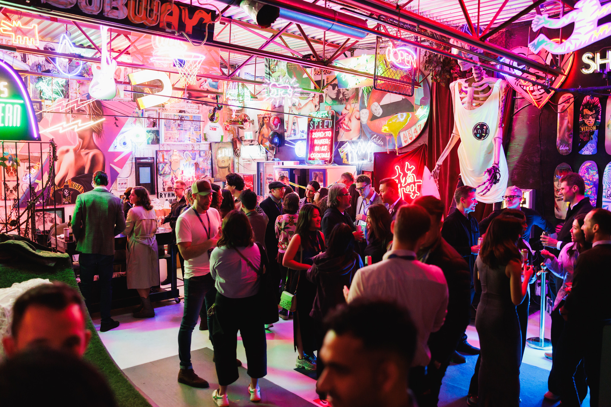A crowd of guests talking and drinking in a warehouse space packed floor to ceiling with neon signs and pop art.