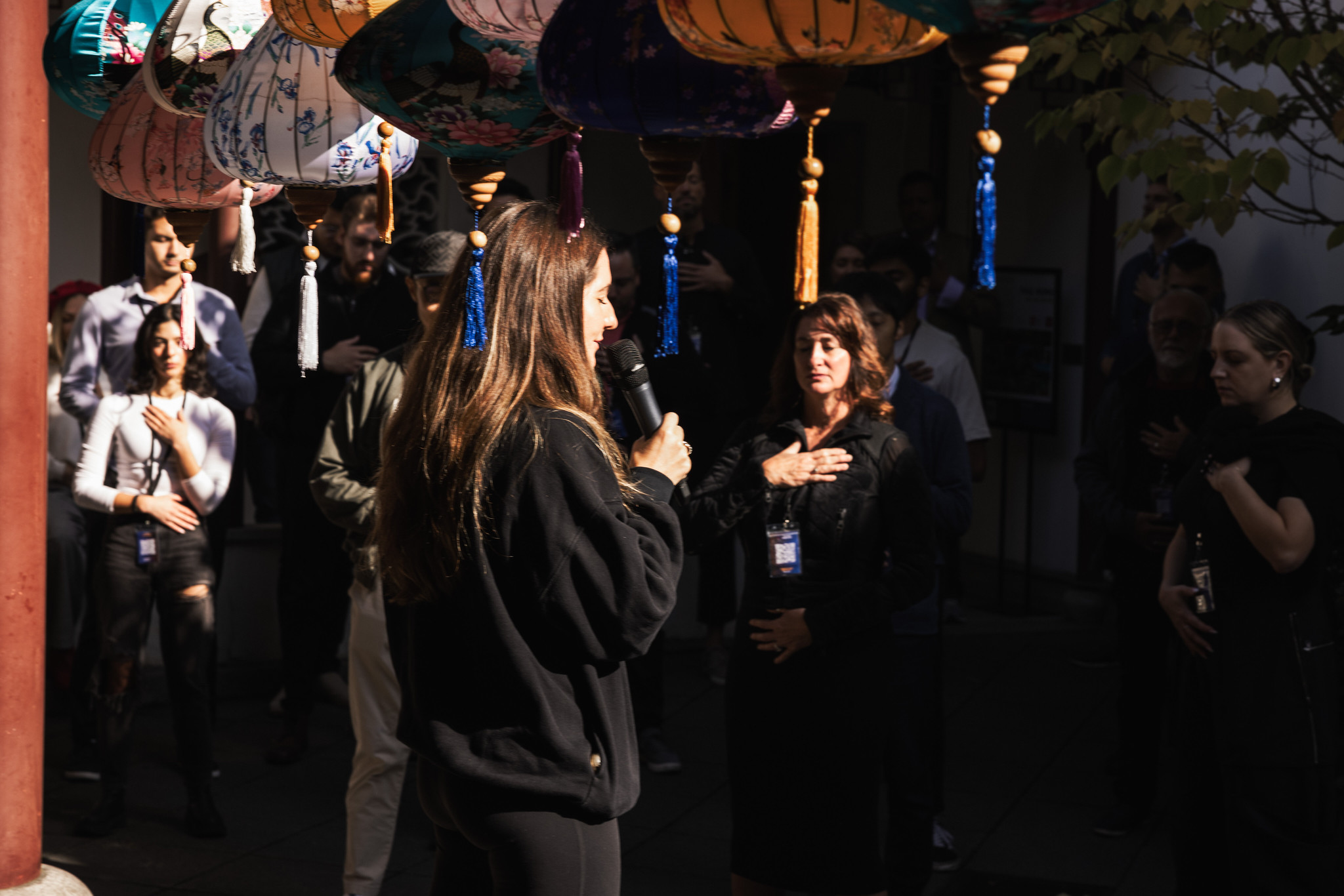 A facilitator leads a group in a guided breathing exercise beneath hanging silk lanterns, participants resting a hand on their chest.