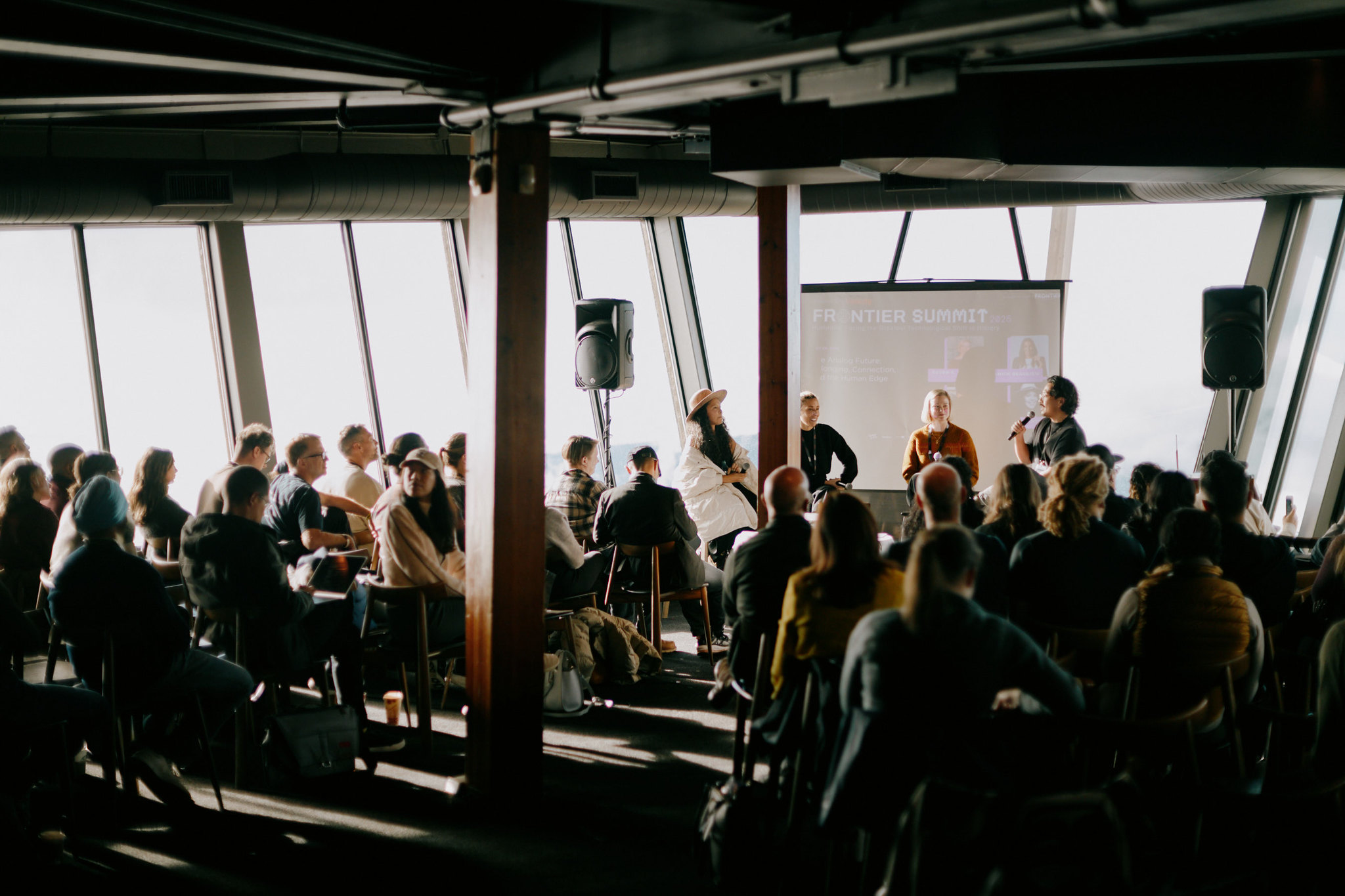 Attendees seated in rows watching three panellists speak at a waterfront venue with floor-to-ceiling windows.