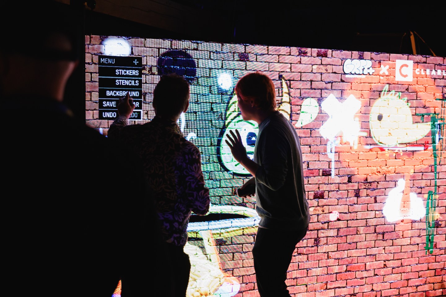 Two attendees paint on an interactive projected brick wall in a darkened room, glowing artwork surrounding them.