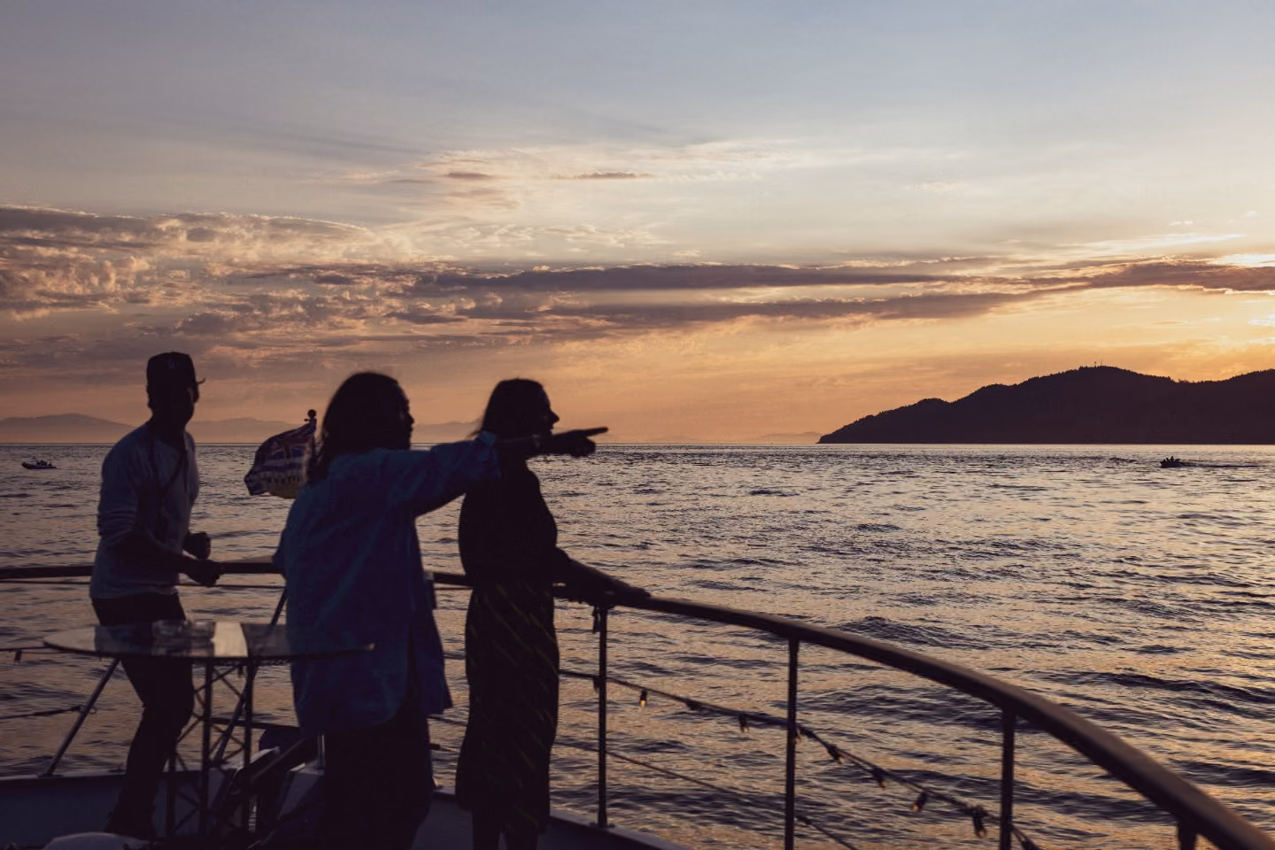 Three people silhouetted on a boat deck at sunset, one pointing across the water toward distant mountains.