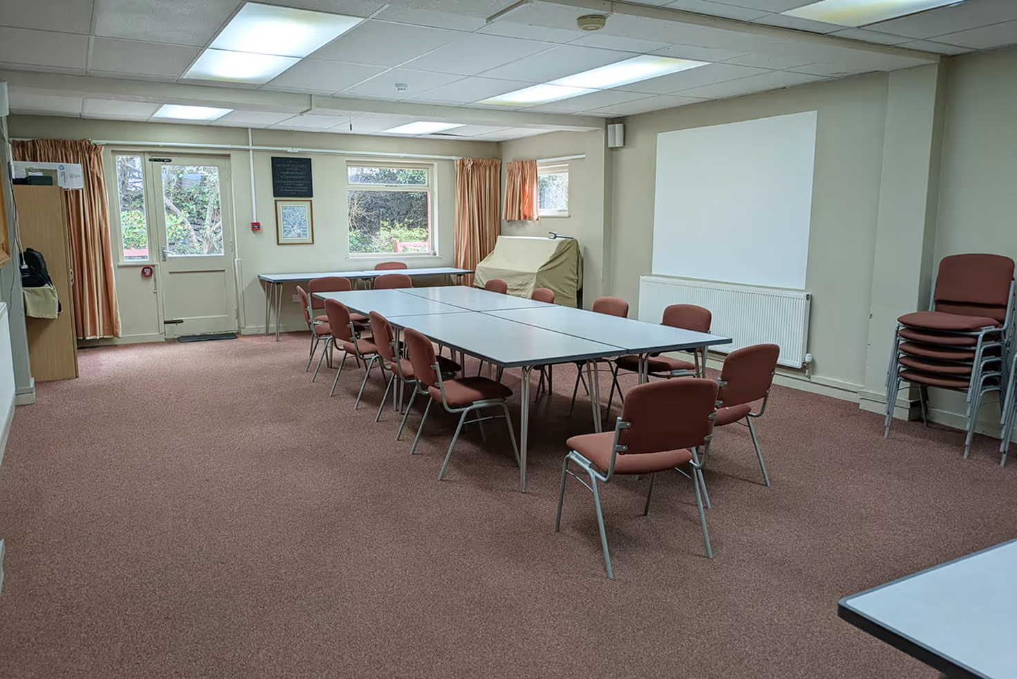 Empty meeting room with rectangular tables arranged in a square and surrounded by brown chairs on carpeted floor.
